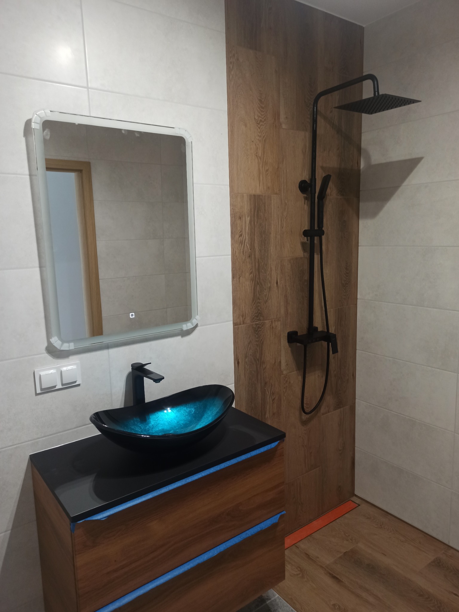 Nowoczesna łazienka z umywalką w kształcie misy o niebieskim wnętrzu, czarną armaturą i deszczownicą, wykończona płytkami imitującymi drewno oraz białymi płytkami ceramicznymi na ścianach. Taśma...