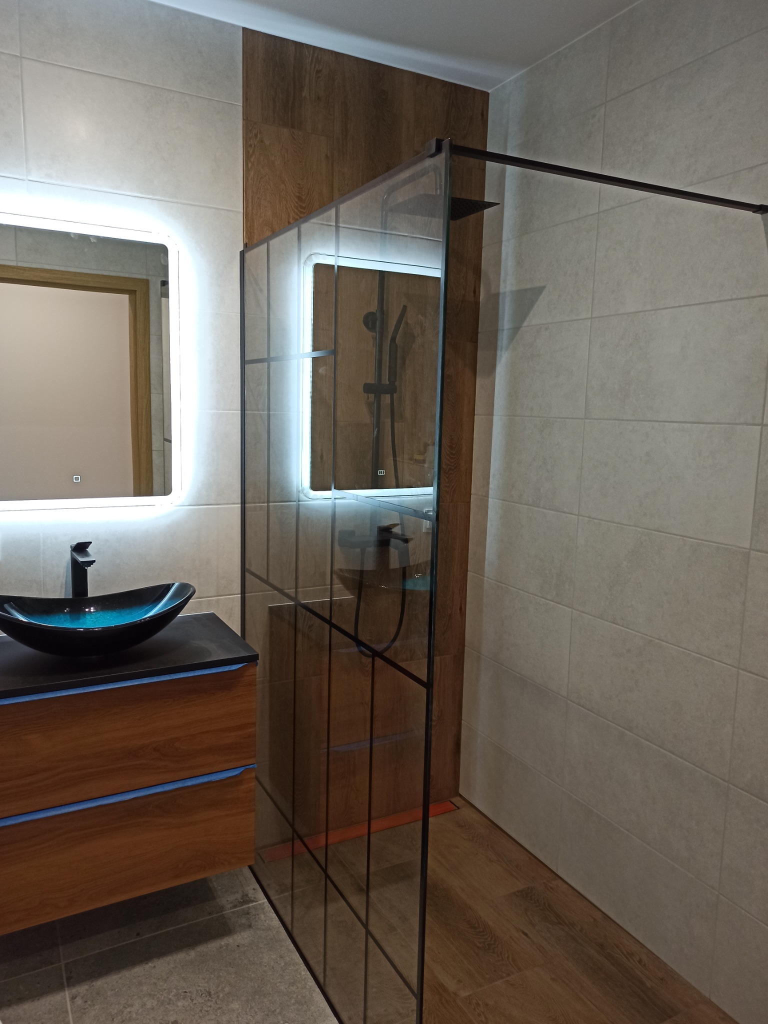 Nowoczesna łazienka z umywalką w kształcie łodzi, podświetlanym lustrem i szklaną kabiną prysznicową z czarnymi profilami, drewnopodobne panele na ścianie i podłodze.