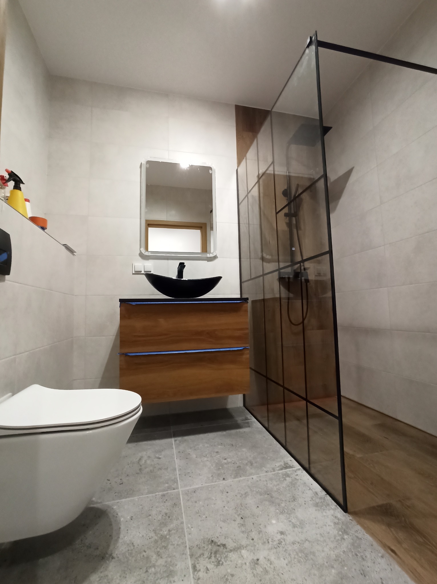 Nowoczesna łazienka z kabiną prysznicową typu walk-in z czarnymi profilami, wiszącą szafką z umywalką w kształcie łodzi oraz toaletą podwieszaną.