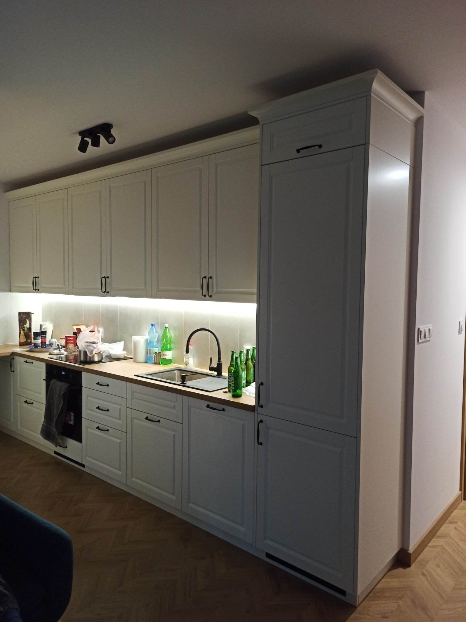 Biała zabudowa kuchenna z oświetleniem podszafkowym, czarną armaturą i panelami podłogowymi ułożonymi w jodełkę; na blacie widoczne butelki i akcesoria kuchenne.