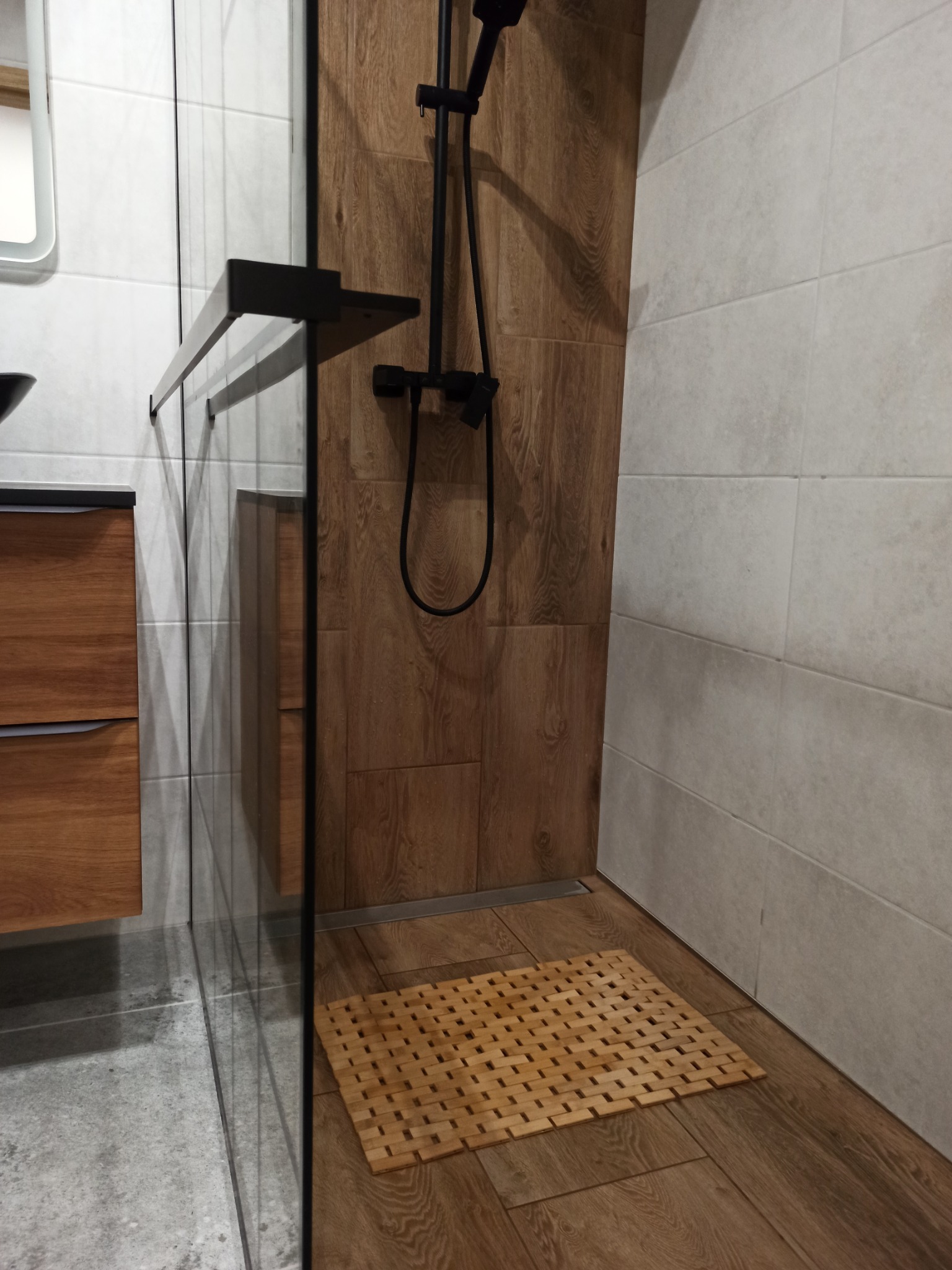 Nowoczesna kabina prysznicowa typu walk-in z czarną armaturą, drewnopodobnymi płytkami na ścianie i podłodze, oraz bambusową wycieraczką.