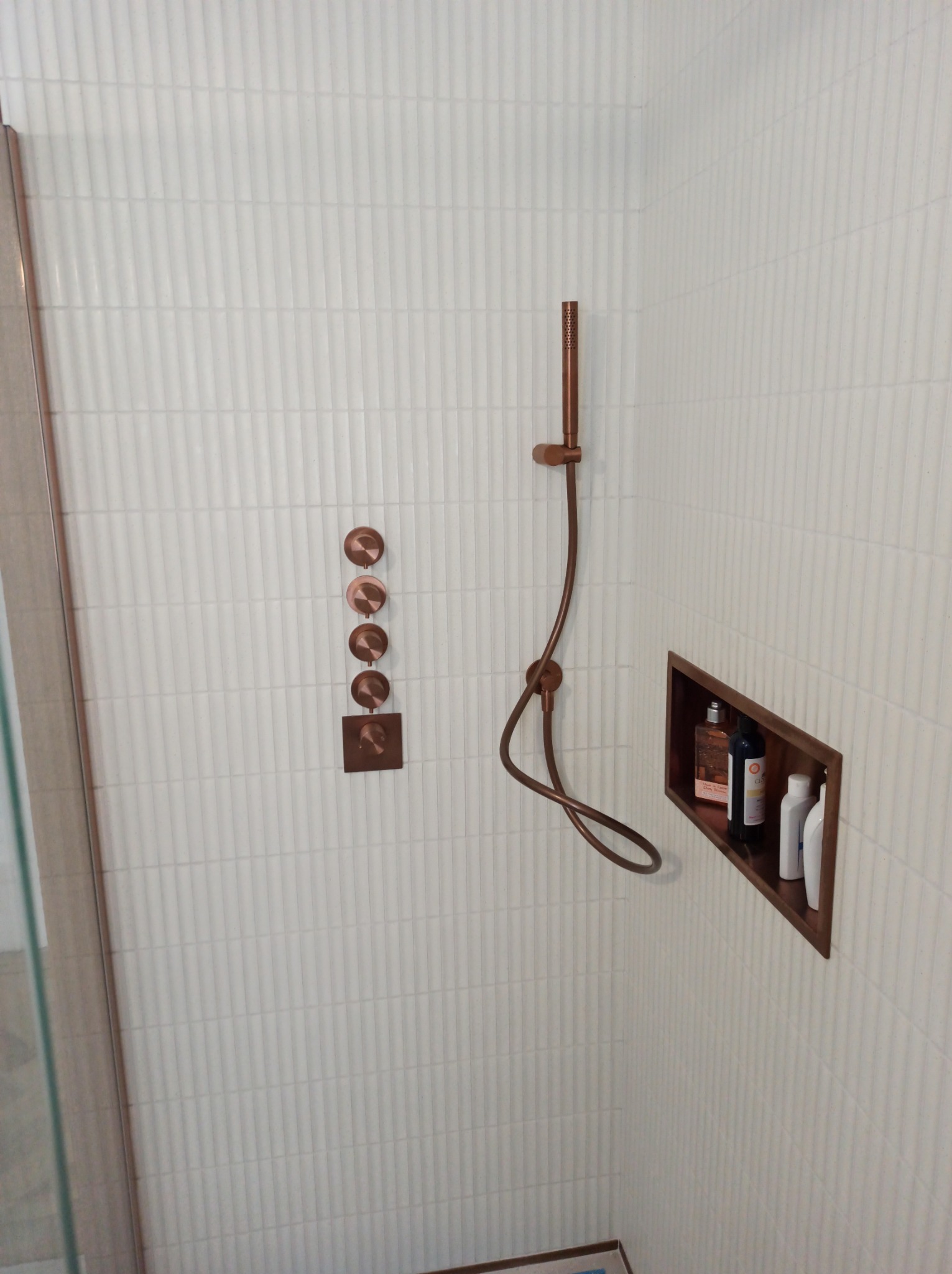 Nowoczesna kabina prysznicowa z białymi, pionowymi płytkami, miedzianą armaturą i wnęką z kosmetykami.