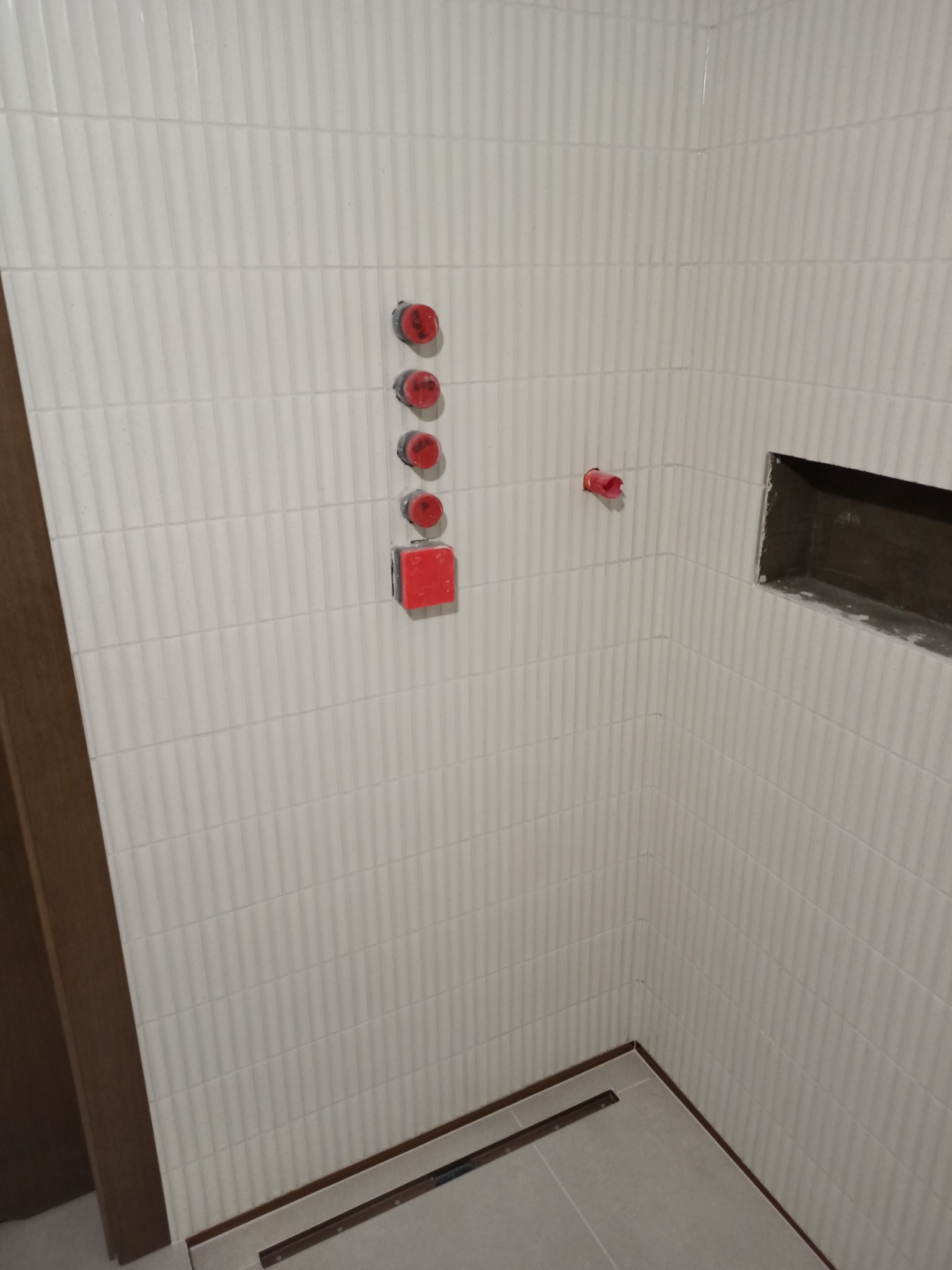 Ściana łazienki wyłożona białymi, wąskimi płytkami z pionowym żłobieniem, widoczne czerwone zaślepki instalacji wodnej i przygotowane miejsce na półkę w ścianie, oraz odpływ liniowy w podłodze...