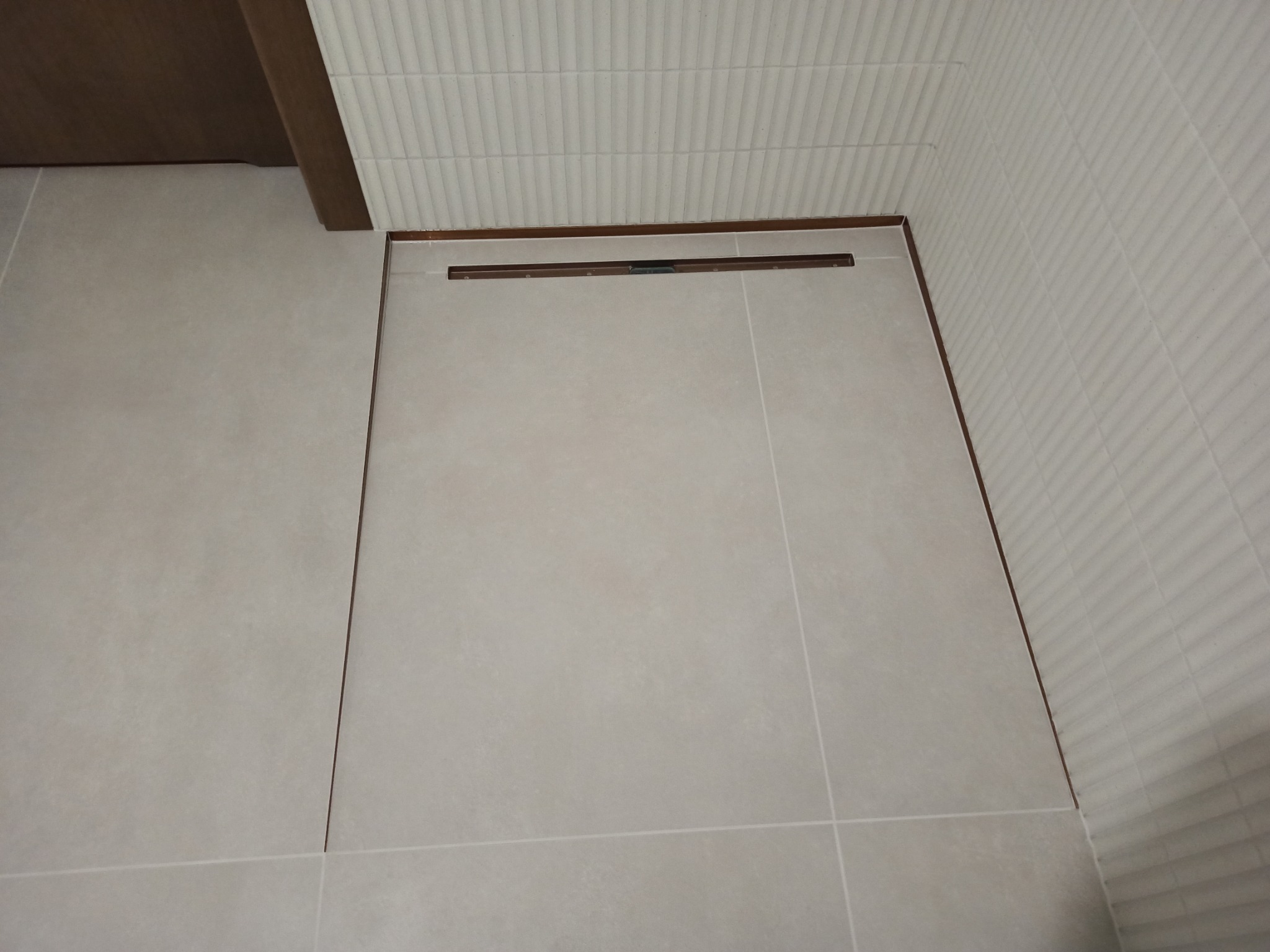 Beżowe płytki podłogowe i ścienne w łazience, z widocznym odpływem liniowym w kolorze brązowym, ułożone w regularny wzór.