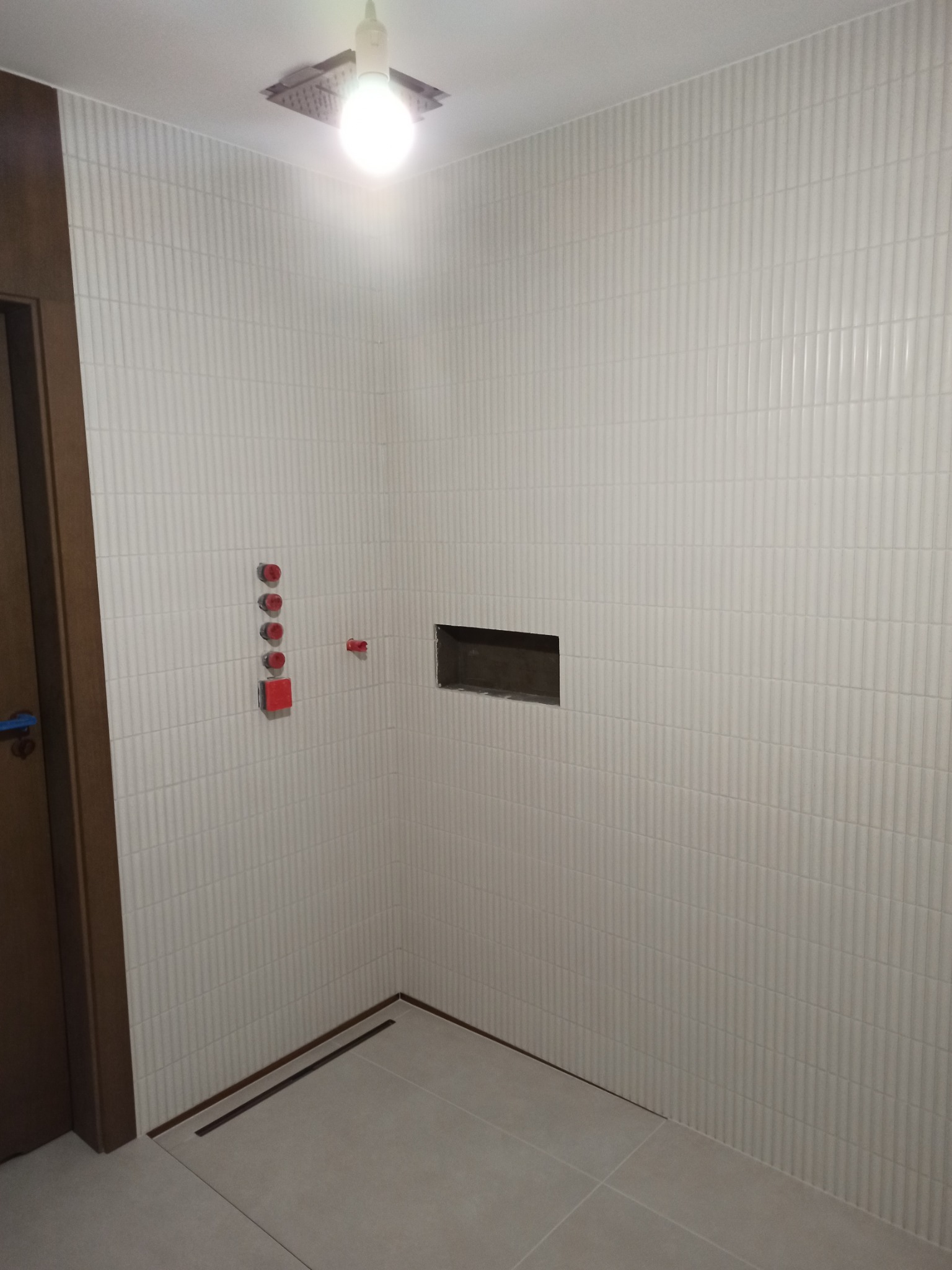 Ściana łazienki wyłożona białymi, pionowo żłobionymi płytkami, z widocznymi elementami instalacji wodnej i otworem rewizyjnym, oraz odpływem liniowym w podłodze.