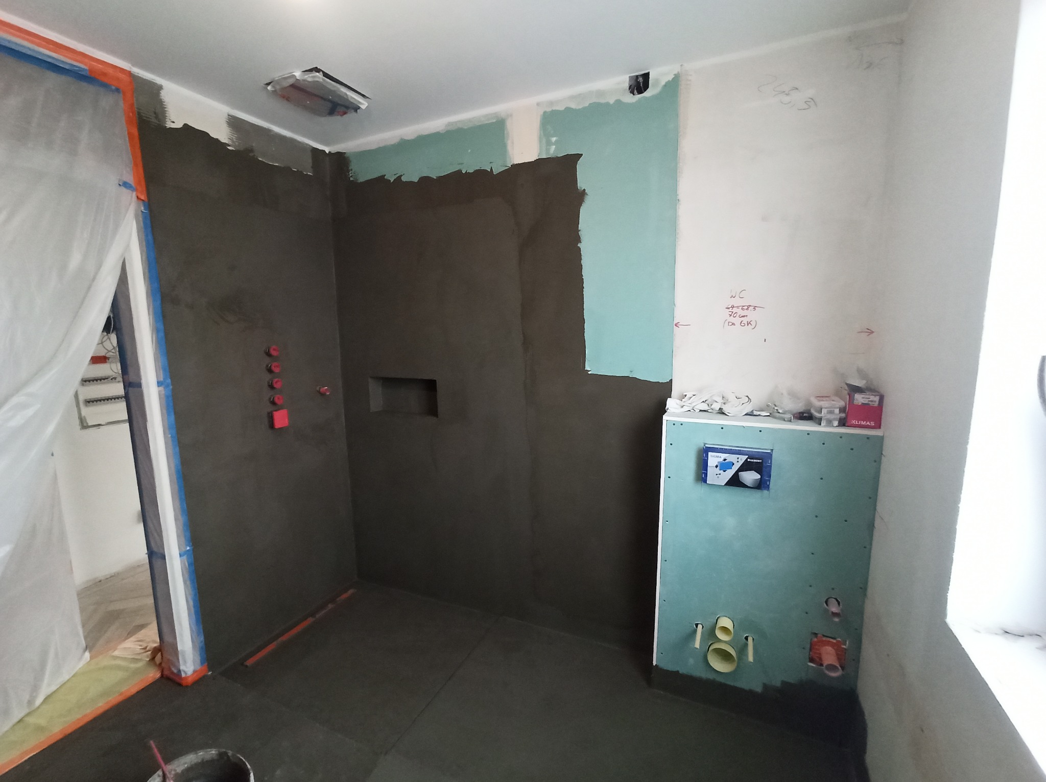 Łazienka w trakcie remontu: ściany pokryte ciemną gładzią szpachlową, częściowo obłożone płytami kartonowo-gipsowymi, widoczna instalacja podtynkowego WC, zabezpieczone folią malarską drzwi.