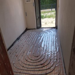 A&M KONSERWACJA 24 SPÓŁKA Z OGRANICZONĄ ODPOWIEDZIALNOŚCIĄ - Białe rury ogrzewania podłogowego ułożone na folii z czerwoną siatką w surowym pomieszczeniu, widok na zewnątrz przez otwarte drzwi.