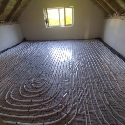 A&M KONSERWACJA 24 SPÓŁKA Z OGRANICZONĄ ODPOWIEDZIALNOŚCIĄ - Białe rury ogrzewania podłogowego rozłożone na folii z czerwoną siatką w surowym pomieszczeniu poddasza, widoczne drewniane belki stropu i okno z zielenią na zewnątrz.