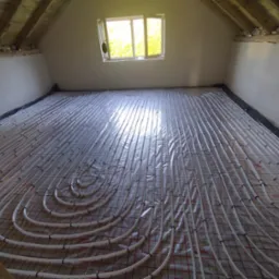 Białe rury ogrzewania podłogowego rozłożone na folii z czerwoną siatką w surowym pomieszczeniu poddasza, widoczne drewniane belki stropu i okno z zielenią na zewnątrz.