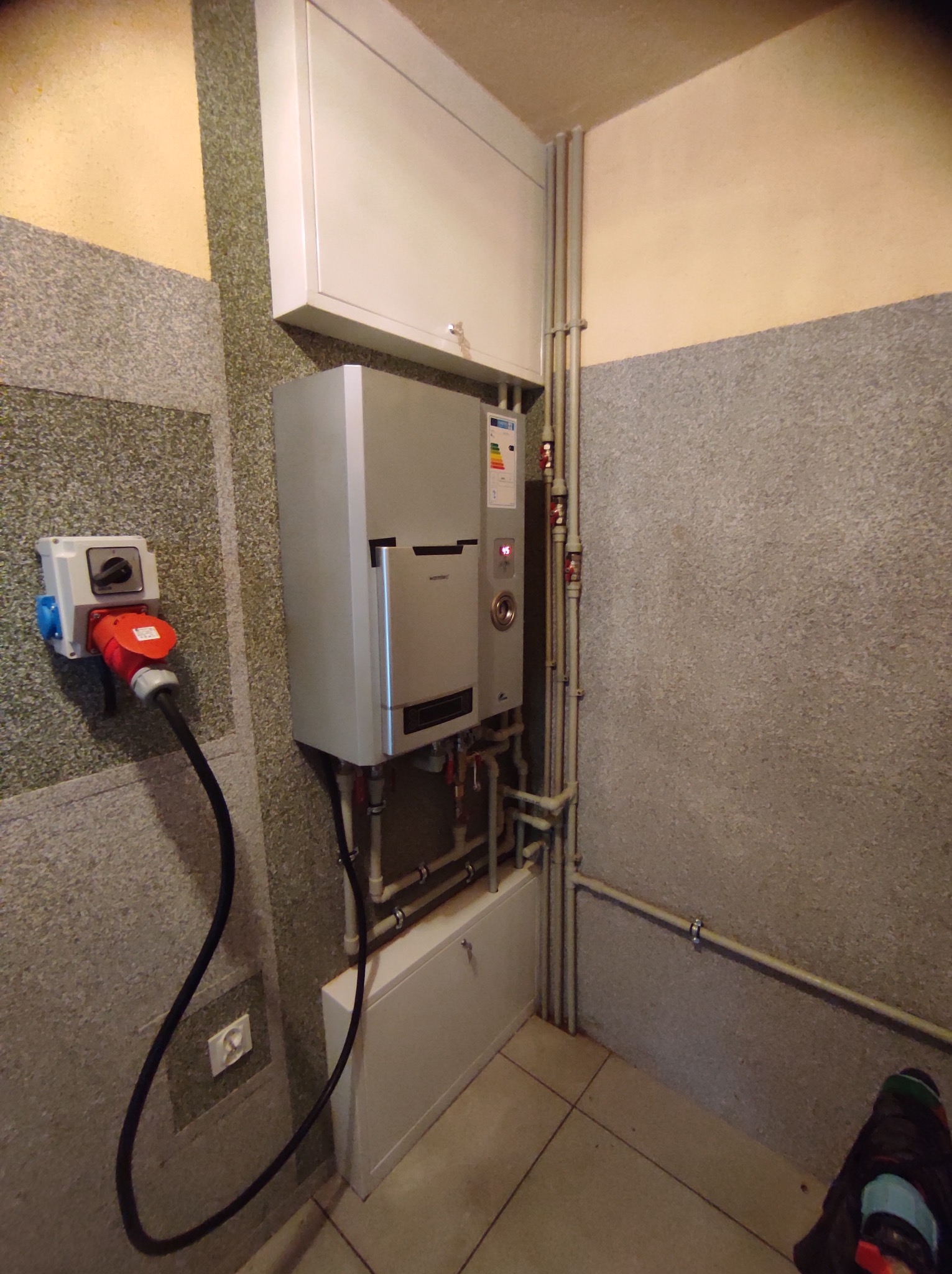 Szary piec grzewczy podłączony do instalacji rurowej, widoczny w pomieszczeniu z szaro-beżowymi ścianami i gniazdem siłowym z wtyczką na ścianie.