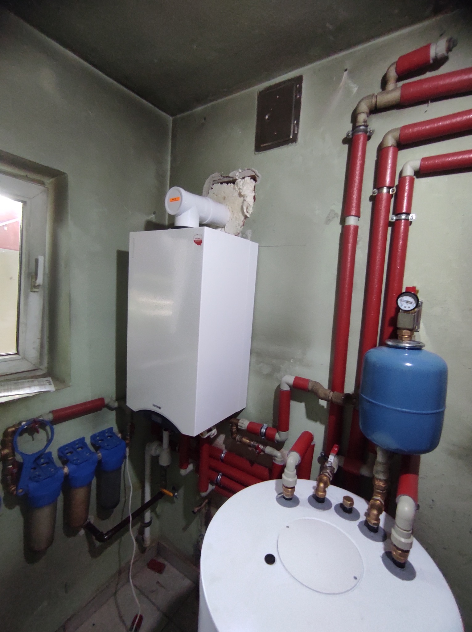 Biały piec gazowy marki Termet zamontowany na ścianie, obok okna i szeregu czerwonych rur grzewczych, z niebieskim zbiornikiem wyrównawczym i filtrami wody, na tle zielonej ściany z widocznymi...