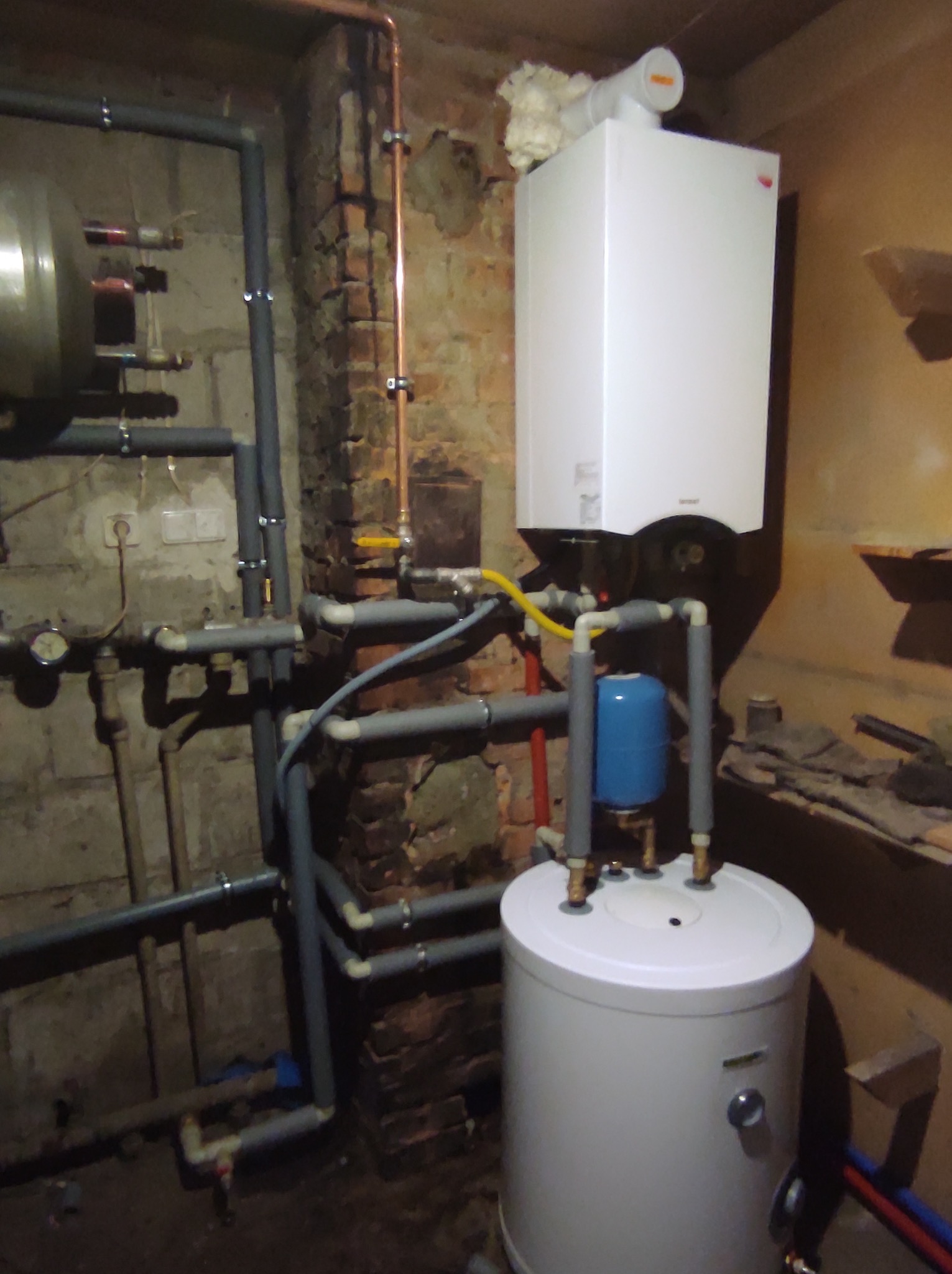 Biały piec gazowy kondensacyjny z podłączonym zasobnikiem ciepłej wody i instalacją hydrauliczną na tle surowej ściany z cegły i betonu.