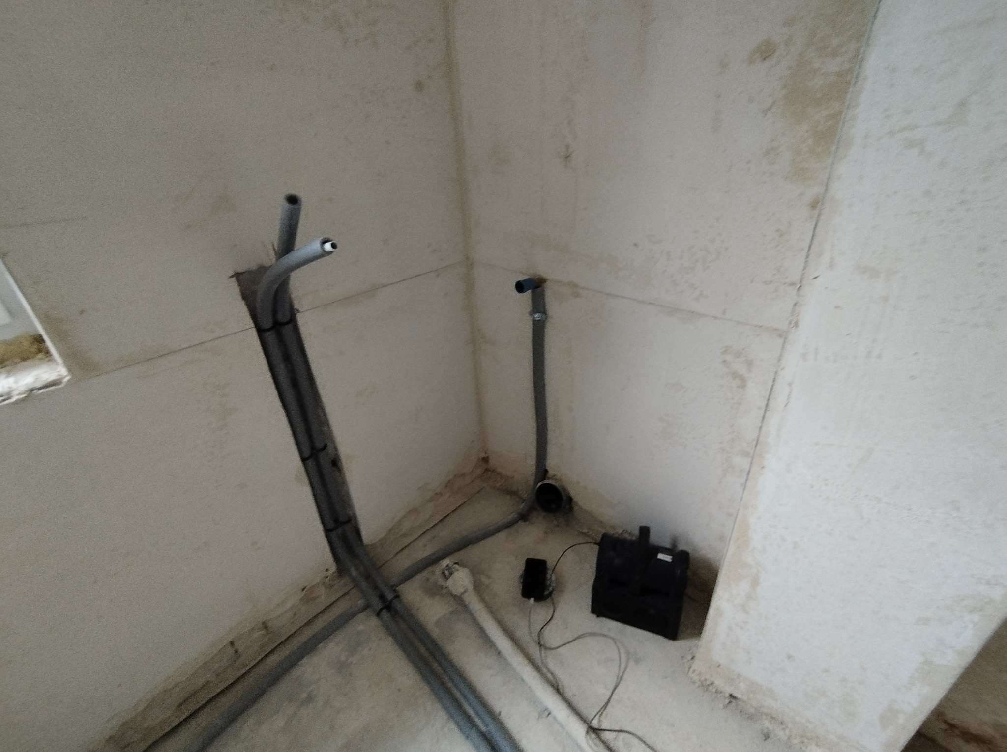 Wnętrze w trakcie prac instalacyjnych: pionowe i poziome szare rury, mały czarny zbiornik oraz biała rura wystająca z podłogi na tle surowych ścian.