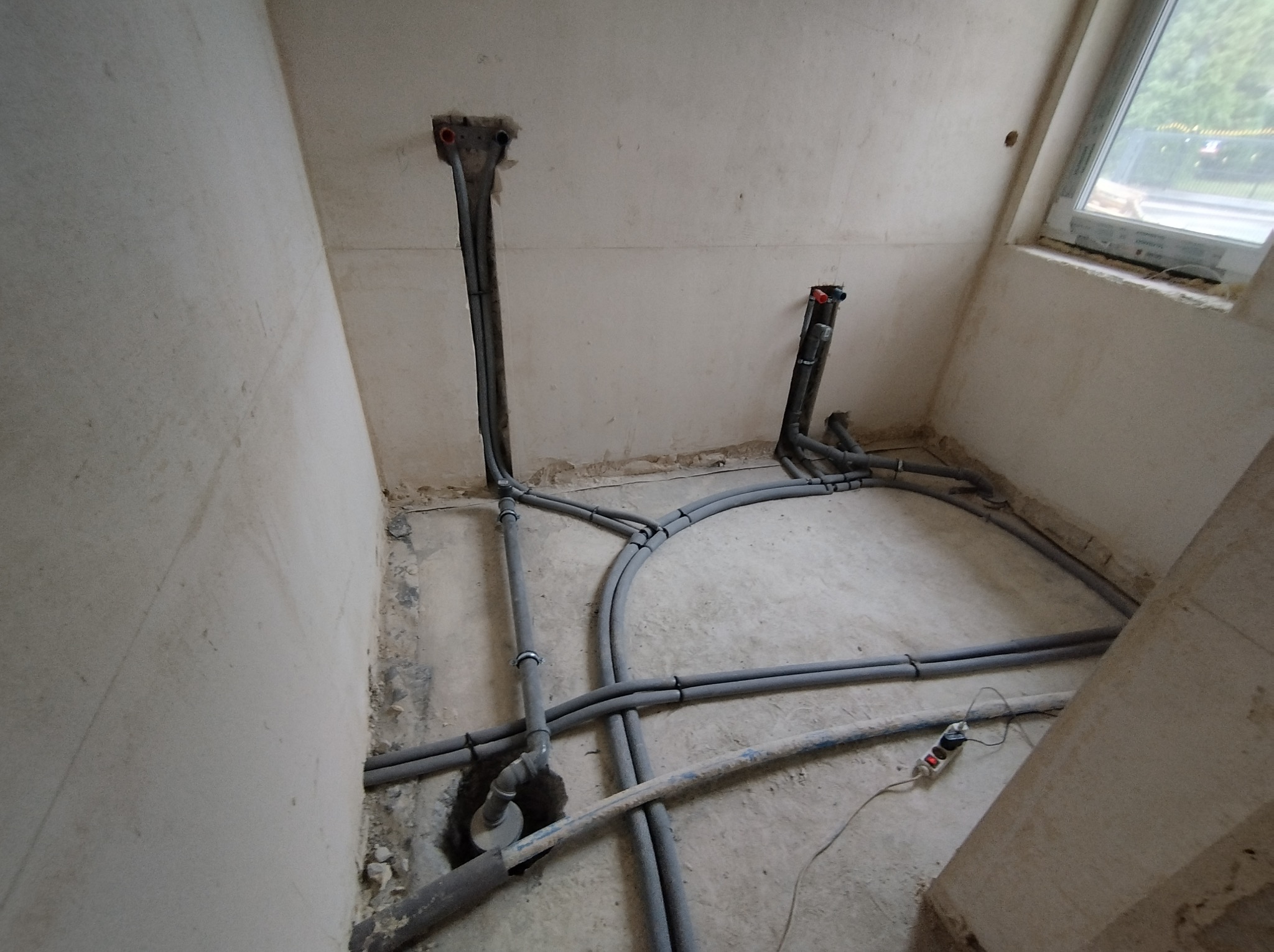 Surowe wnętrze z widocznymi pionami rur instalacji wodnej i grzewczej wychodzącymi ze ścian i podłogi, szare rury prowadzone po podłodze, widoczny przedłużacz z włączonymi diodami.