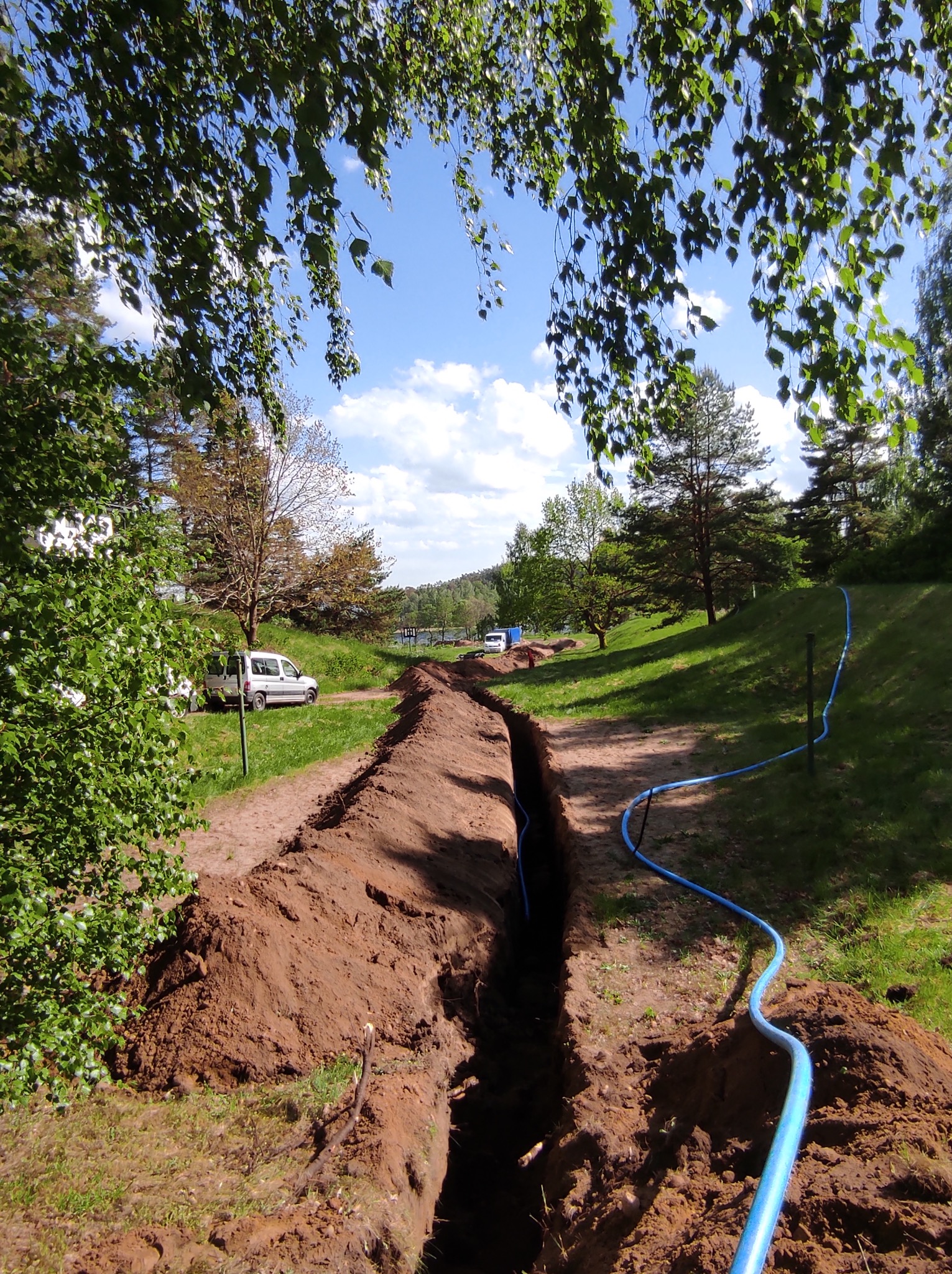 Wykopany rów w ziemi z niebieskim przewodem biegnącym wzdłuż niego, widok z góry, w tle samochody i drzewa