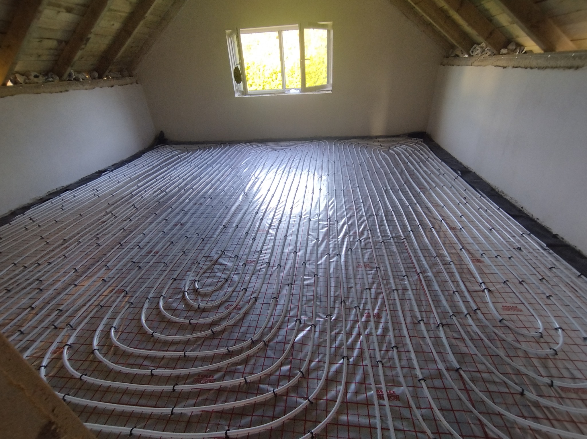 Białe rury ogrzewania podłogowego rozłożone na folii z czerwoną siatką w surowym pomieszczeniu poddasza, widoczne drewniane belki stropu i okno z zielenią na zewnątrz.