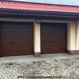 Daniel Jedrzejkiewicz - Dwie zamknięte, segmentowe bramy garażowe w kolorze ciemnego drewna, zamontowane w budynku z beżową elewacją i czerwoną dachówką, oświetlone zewnętrzną lampą.