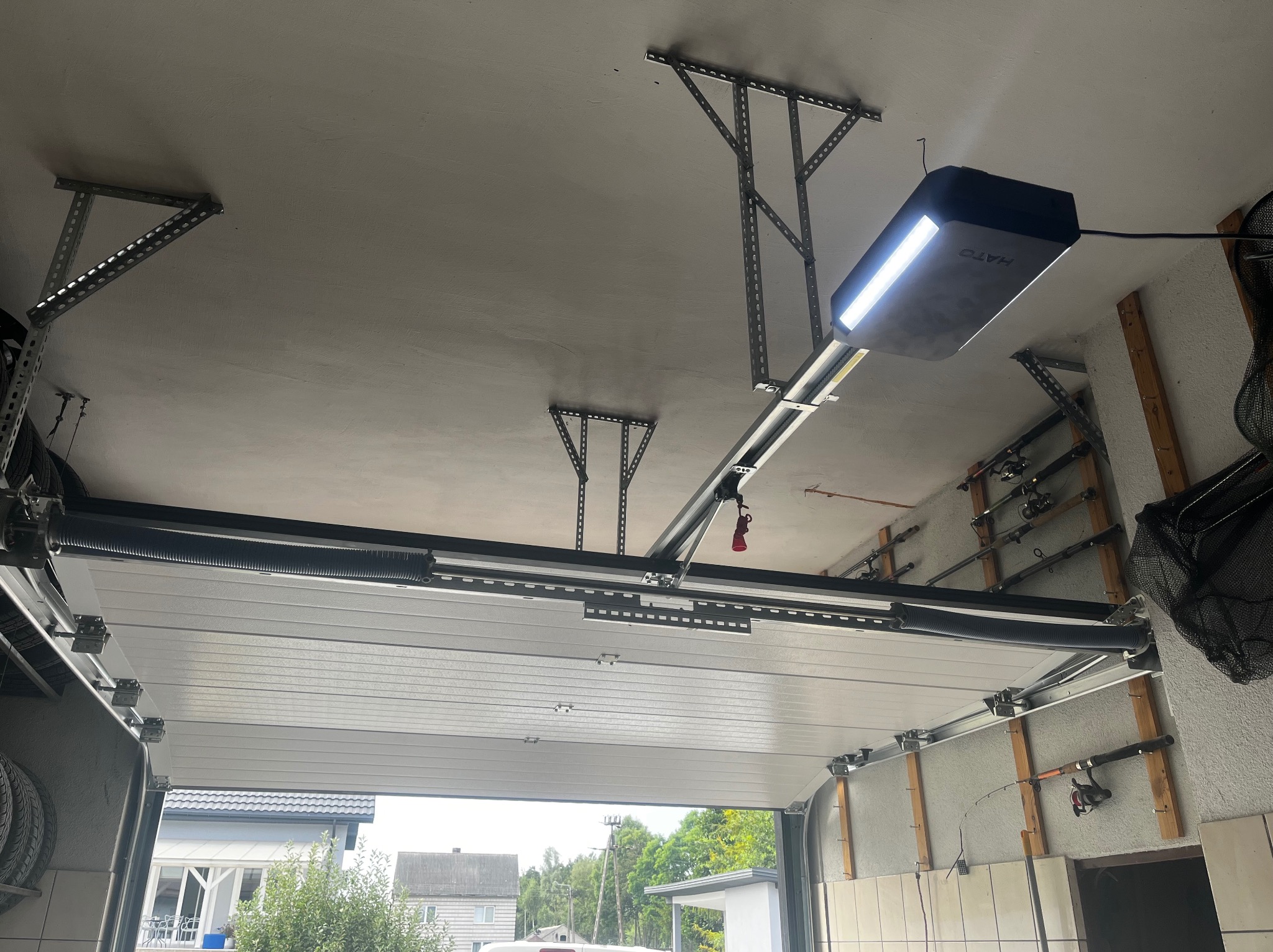 Wnętrze garażu z widocznym mechanizmem otwierania bramy segmentowej, sprężyny skrętne, szyny montażowe, automat HATO z oświetleniem LED, w tle wędki na ścianie.