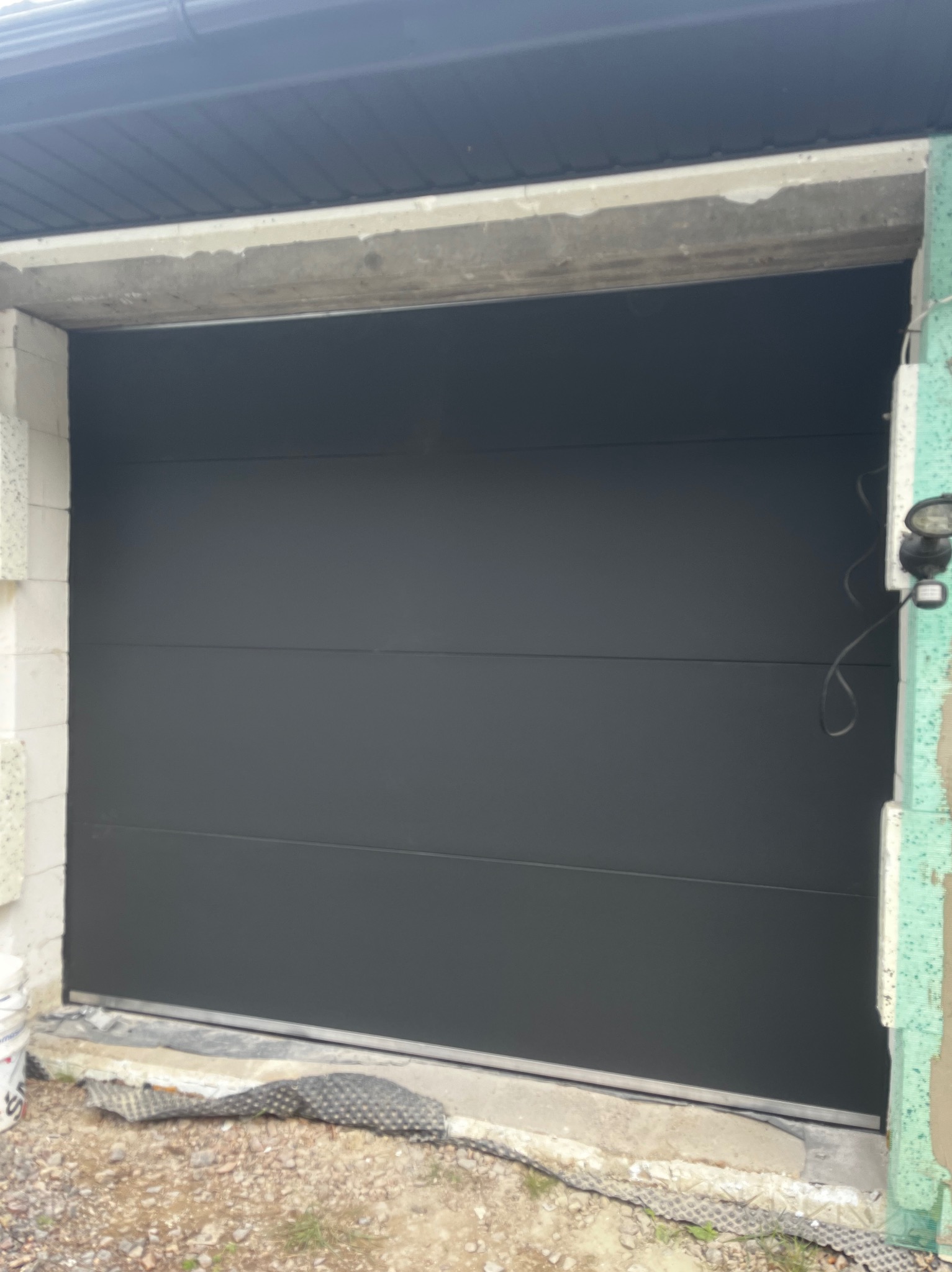 Zamknięta, segmentowa brama garażowa w kolorze antracytowym, widoczna od wewnątrz garażu w trakcie montażu, z surowym wykończeniem ścian i podłogi.