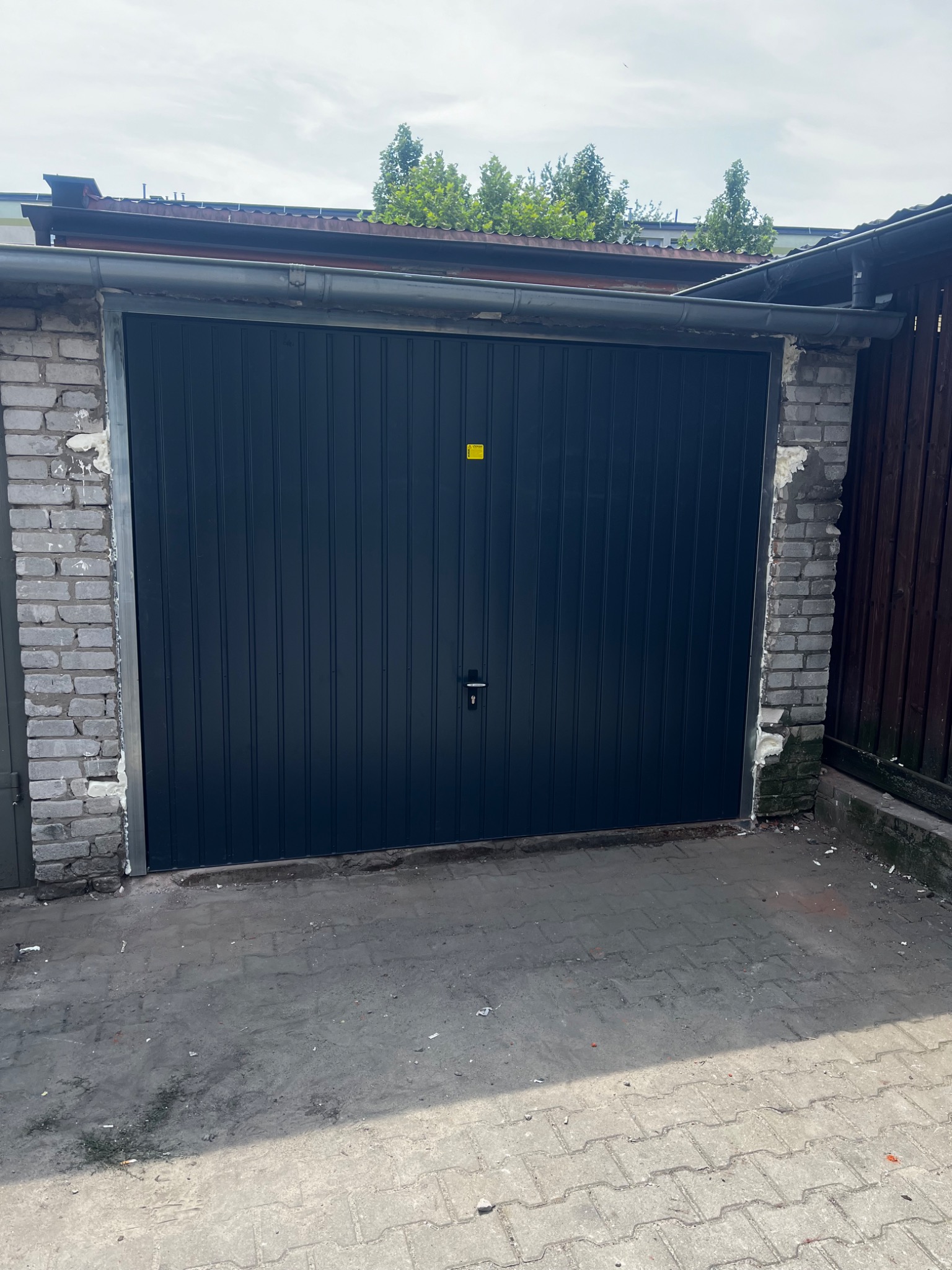 Ciemna, pionowo panelowa brama garażowa w murze z cegły, z widocznymi śladami pianki montażowej i małą żółtą naklejką, na tle betonowego podjazdu i fragmentu drewnianego ogrodzenia.