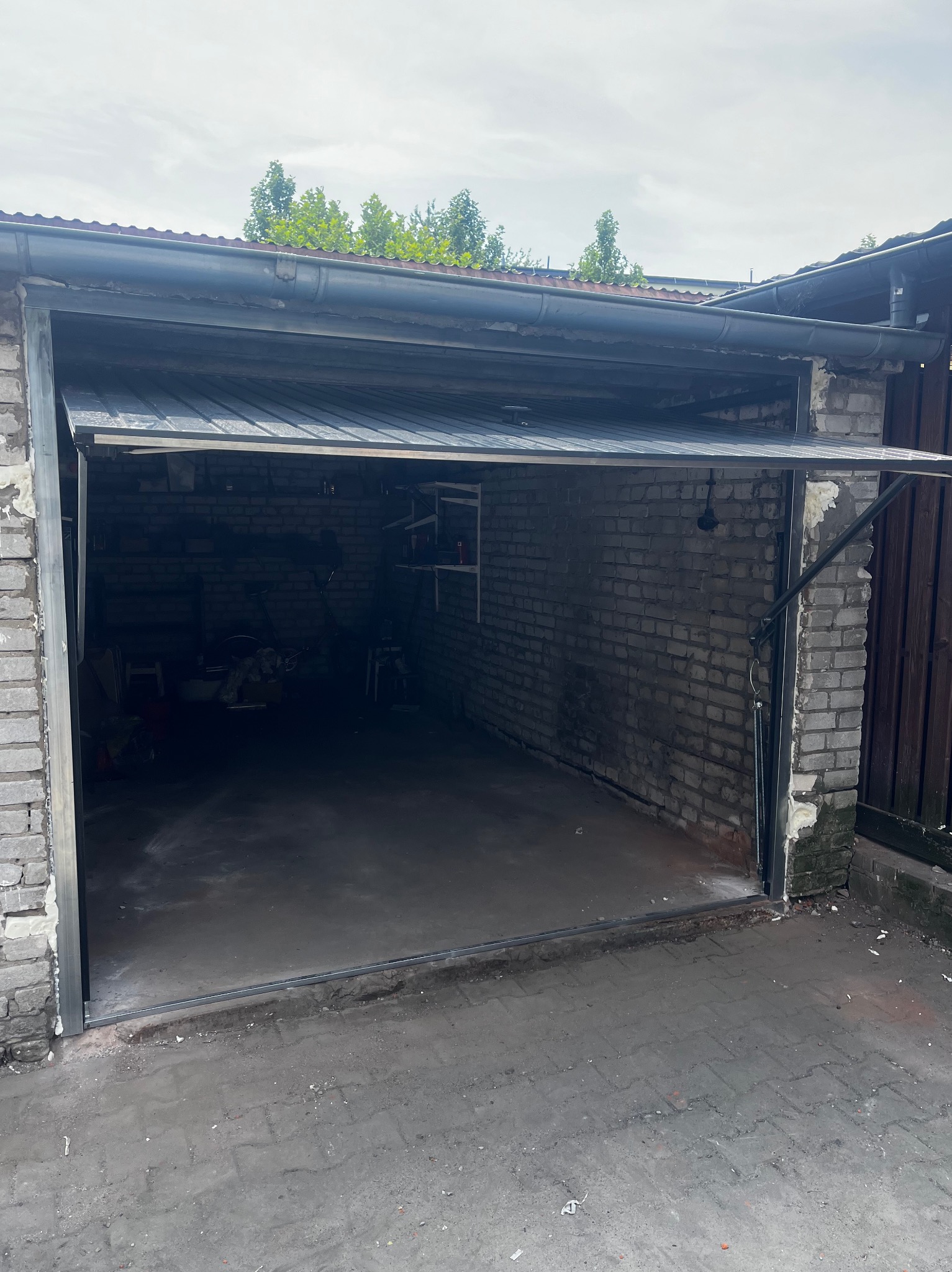 Otwarta brama garażowa segmentowa, widoczna ceglana ściana garażu i betonowa posadzka, brama częściowo podniesiona, widoczne mechanizmy podnoszenia.