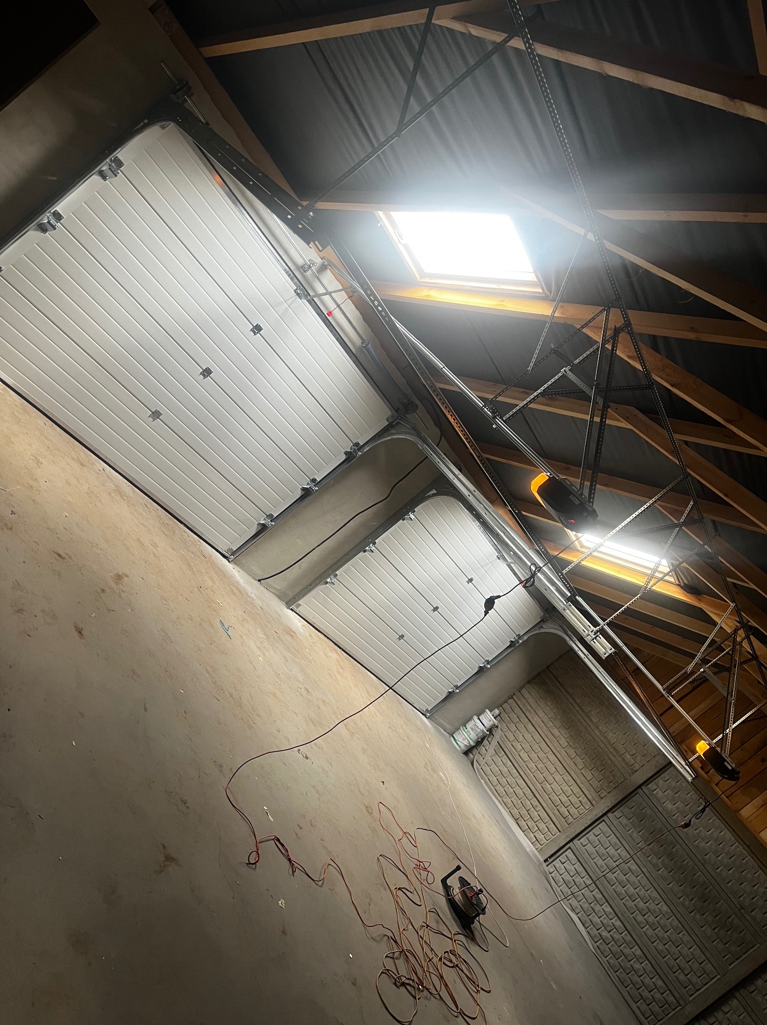 Wnętrze garażu z zamontowaną białą segmentową bramą, widoczne prowadnice, łańcuch i silnik napędu pod sufitem, narzędzia i przewody na podłodze, oświetlenie górne ze świetlika.