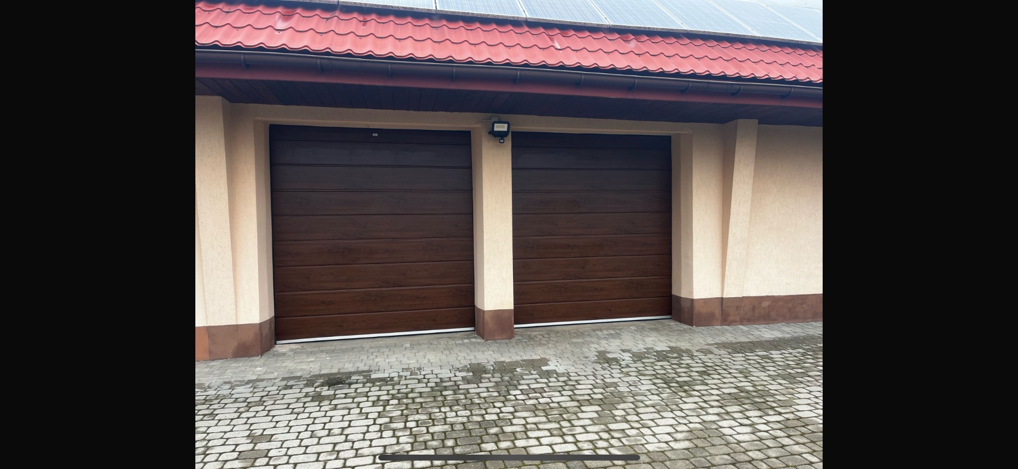 Dwie zamknięte, segmentowe bramy garażowe w kolorze ciemnego drewna, zamontowane w budynku z beżową elewacją i czerwoną dachówką, oświetlone zewnętrzną lampą.