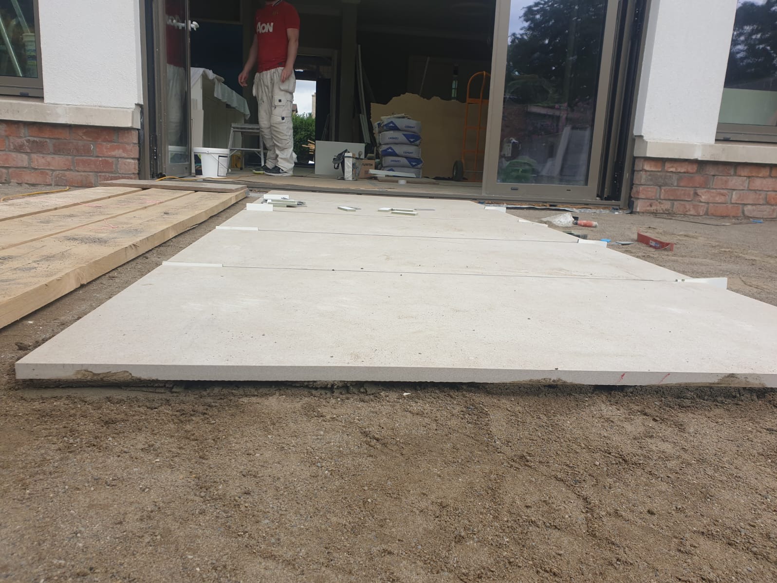 Układanie jasnych płyt na podłożu piaskowym, widoczne narzędzia i materiały budowlane. Prace wykończeniowe przy tarasie z dużymi oknami.