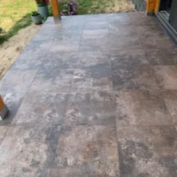 Wyłożony płytkami taras przy drewnianym domu z widocznymi donicami z kwiatami, ziemią i trawą w tle.