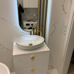 PavBud - Nowoczesna łazienka z umywalką nablatową na białej szafce z dwoma szufladami i złotymi uchwytami, złota bateria, lustro z podświetleniem LED i elementami dekoracyjnymi w kolorze złotym i czarnym...