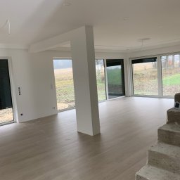 PavBud - Jasne, przestronne wnętrze w trakcie wykańczania, z dużymi oknami z roletami, świeżo pomalowanymi ścianami, drewnopodobną podłogą i surowymi, betonowymi schodami.