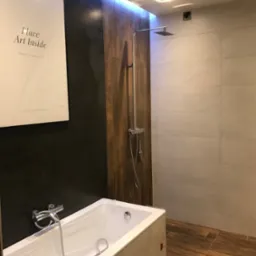 Nowoczesna łazienka z wanną obłożoną płytkami, panelami imitującymi drewno i deszczownicą; na ścianie plakat z napisem 'Place Art Inside'.