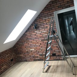 PavBud - Poddasze po remoncie: ceglana ściana z oknem dachowym, aluminiowa drabina, jasna drewniana podłoga, czarne oświetlenie punktowe.
