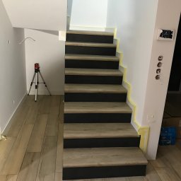 PavBud - Schody z drewnopodobnymi stopniami i czarnymi podstopnicami, zabezpieczone taśmą malarską, z laserowym niwelatorem na trójnogu w tle oraz widocznymi otworami na gniazdka elektryczne.
