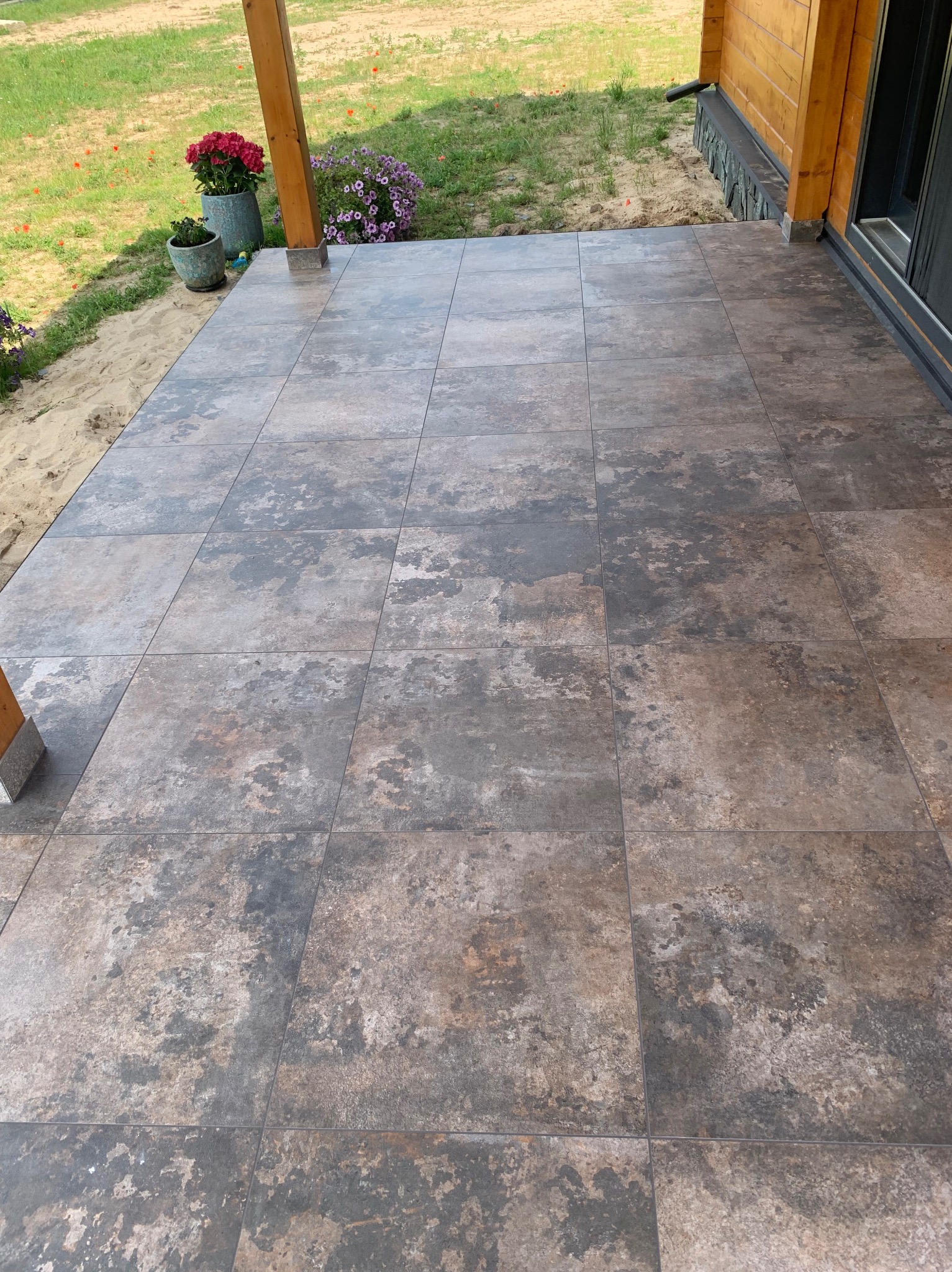 Wyłożony płytkami taras przy drewnianym domu z widocznymi donicami z kwiatami, ziemią i trawą w tle.