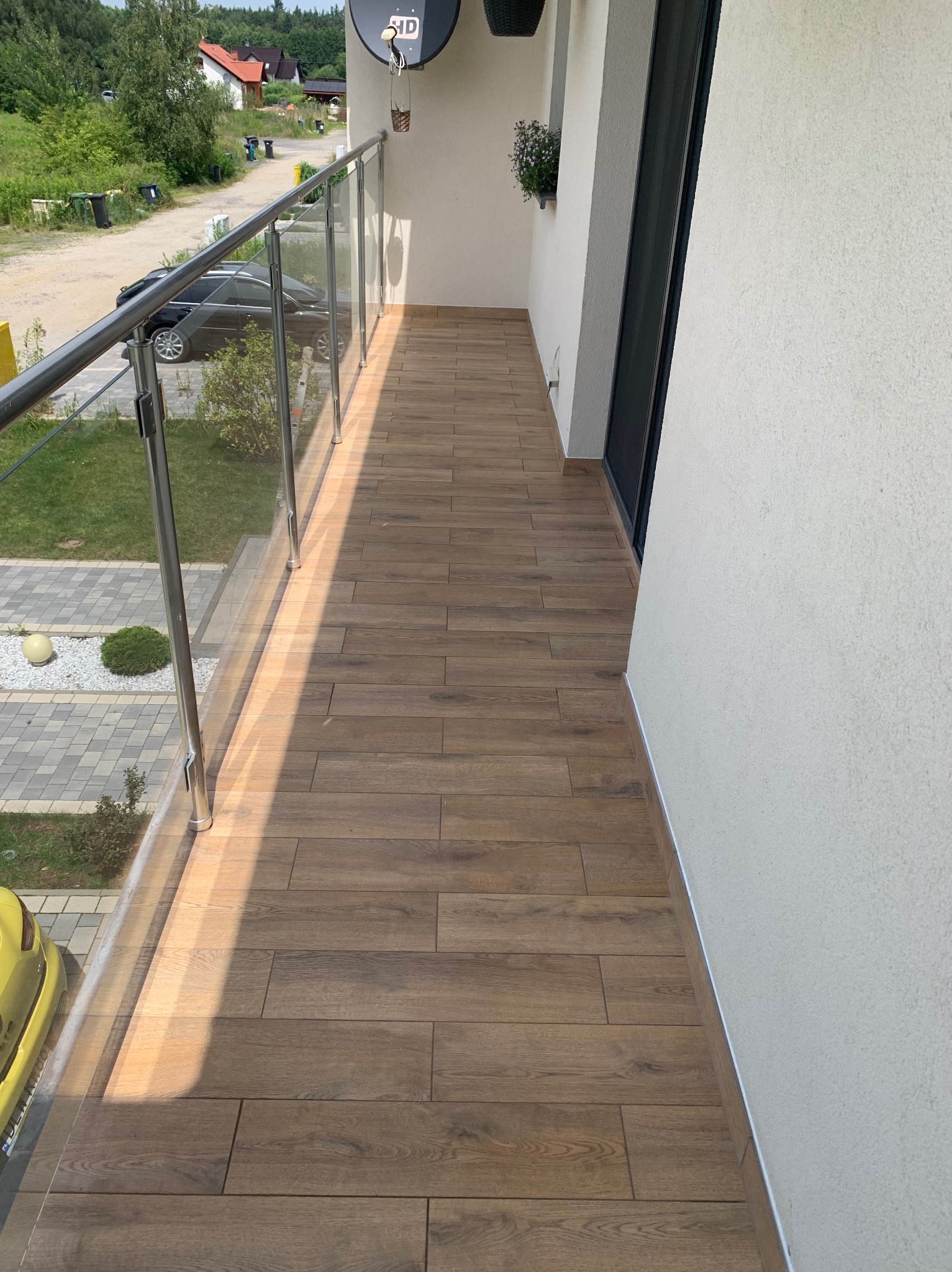 Wykończony balkon z drewnopodobną terakotą, szklaną balustradą i widokiem na okolicę.