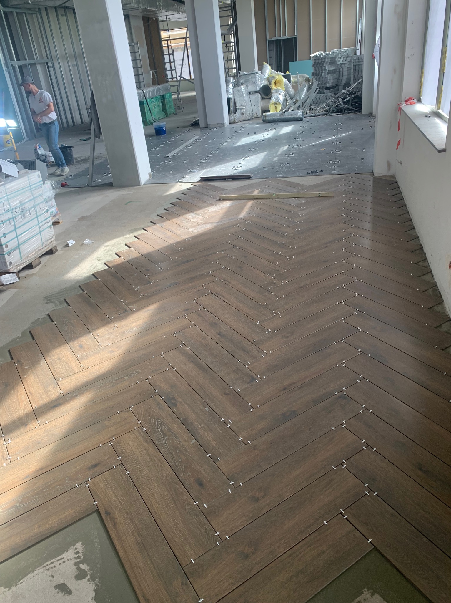 Układanie brązowych płytek podłogowych w jodełkę, widoczne krzyżyki dystansowe i poziomica, w tle remontowany lokal z metalowymi profilami ścian i oknami.