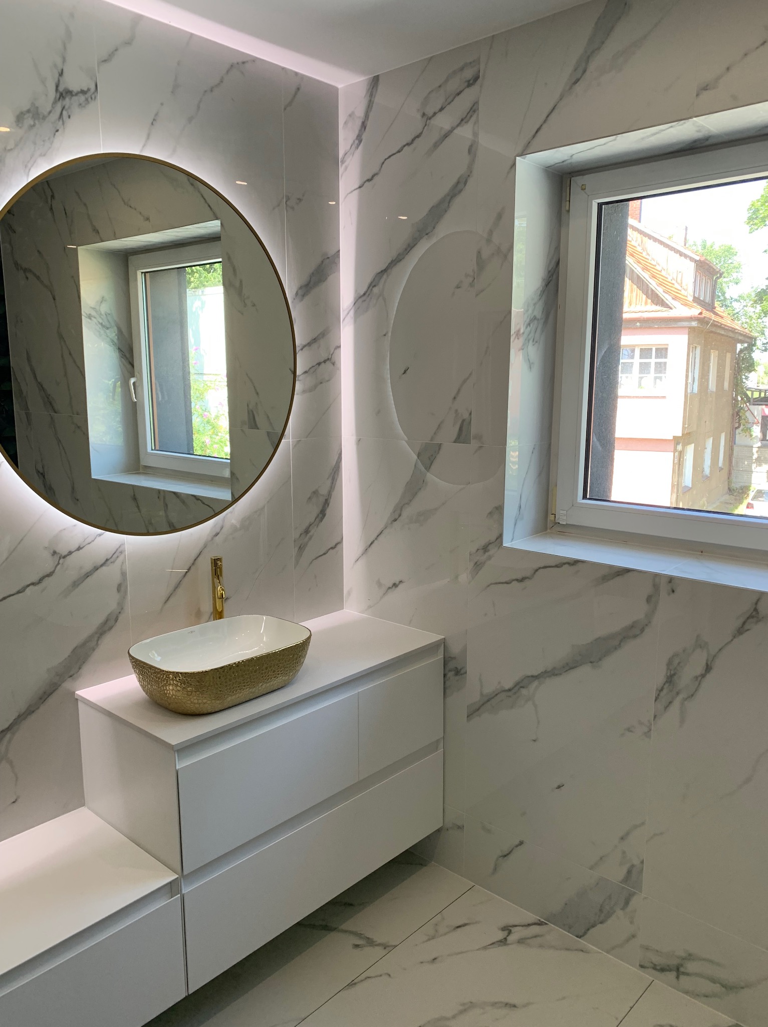 Elegancka łazienka z białymi płytkami imitującymi marmur, okrągłym lustrem z podświetleniem LED, złotą umywalką i minimalistyczną białą szafką pod umywalką, widok z okna na budynek.