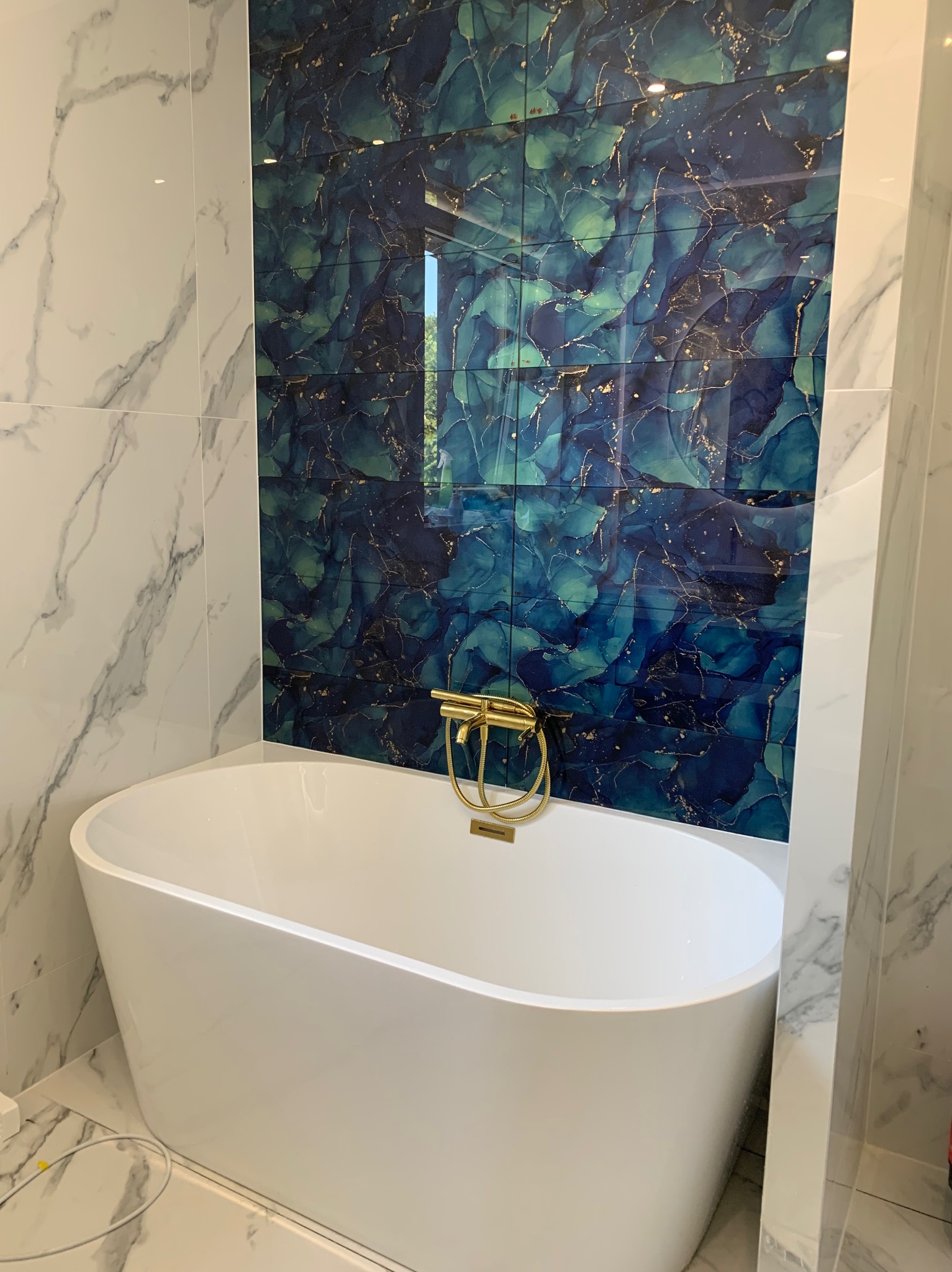 Nowoczesna łazienka z wolnostojącą białą wanną, baterią w kolorze złotym i ścianą wyłożoną płytkami w odcieniach niebieskiego i zieleni z marmurkowym wzorem. Druga ściana wyłożona płytkami...