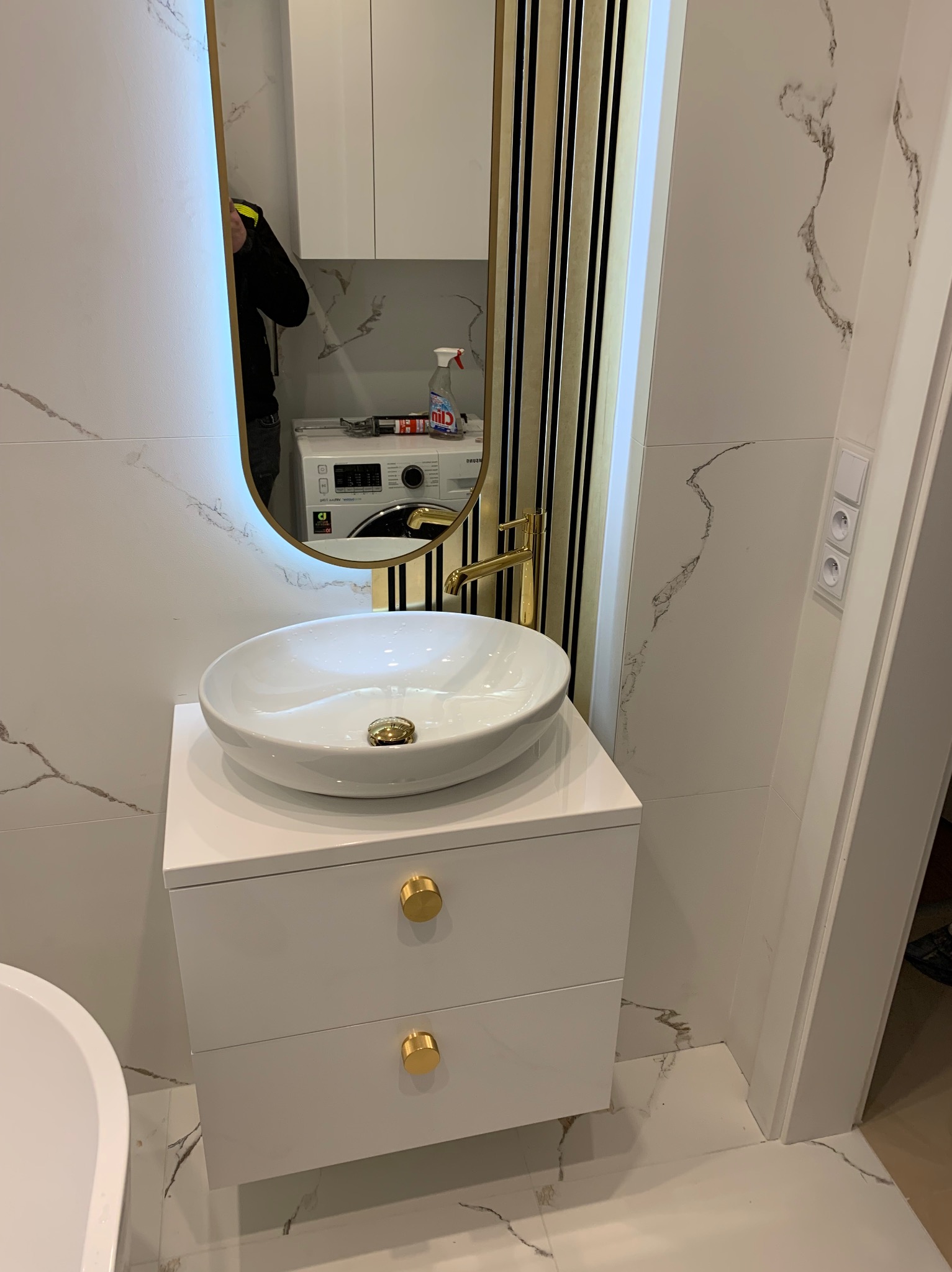 Nowoczesna łazienka z umywalką nablatową na białej szafce z dwoma szufladami i złotymi uchwytami, złota bateria, lustro z podświetleniem LED i elementami dekoracyjnymi w kolorze złotym i czarnym...