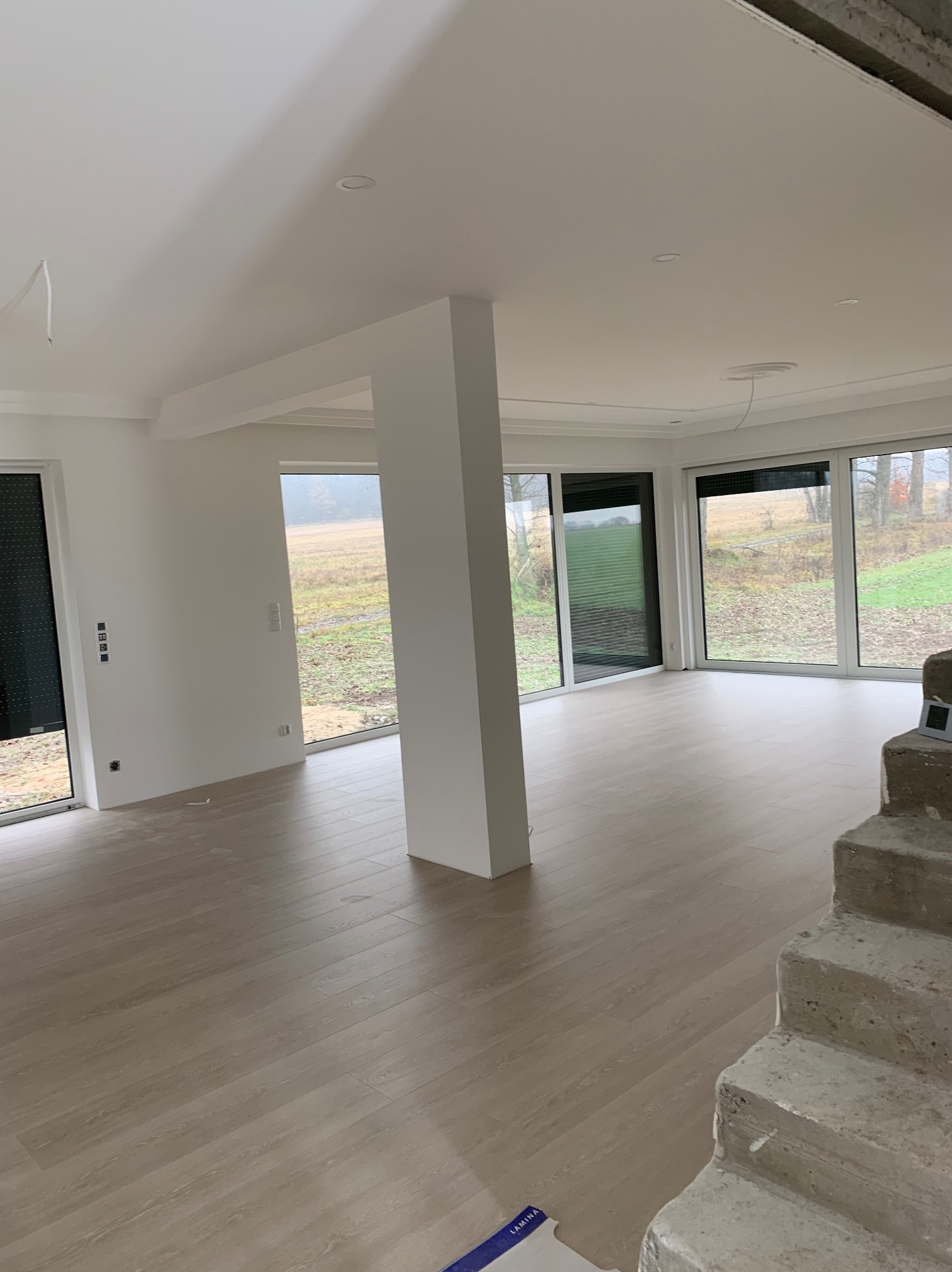 Jasne, przestronne wnętrze w trakcie wykańczania, z dużymi oknami z roletami, świeżo pomalowanymi ścianami, drewnopodobną podłogą i surowymi, betonowymi schodami.