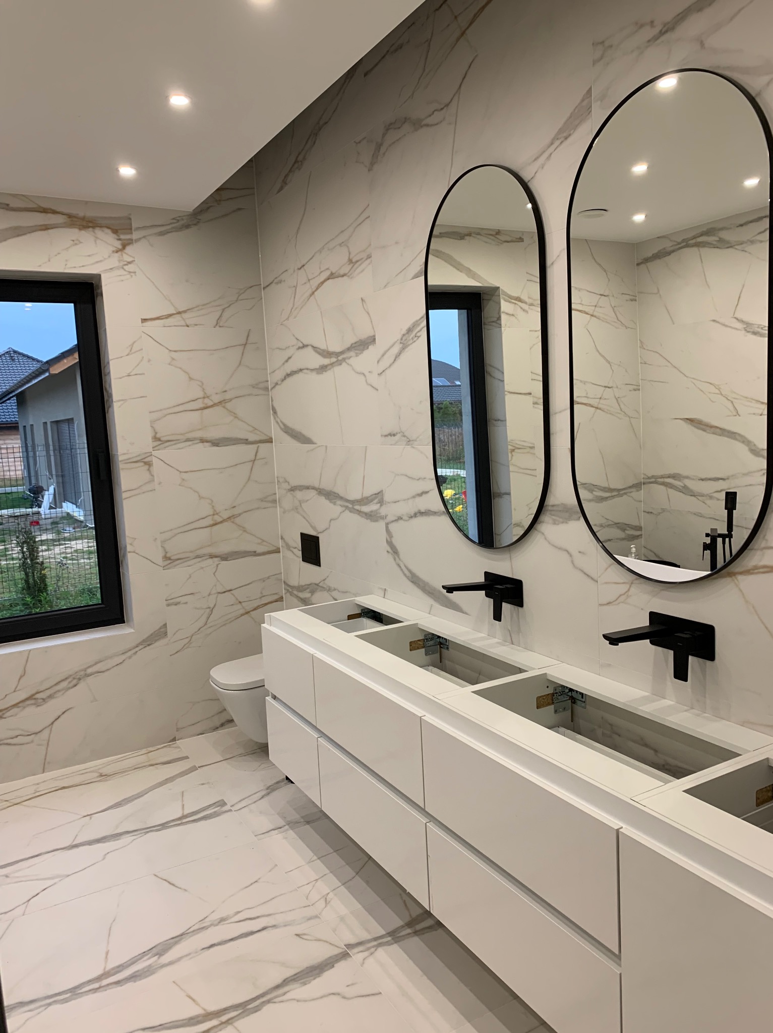 Nowoczesna łazienka w trakcie wykańczania: białe marmurowe płytki na ścianach i podłodze, podwójna biała szafka pod umywalkę z czarnymi bateriami, dwa owalne lustra w czarnych ramach, widok...