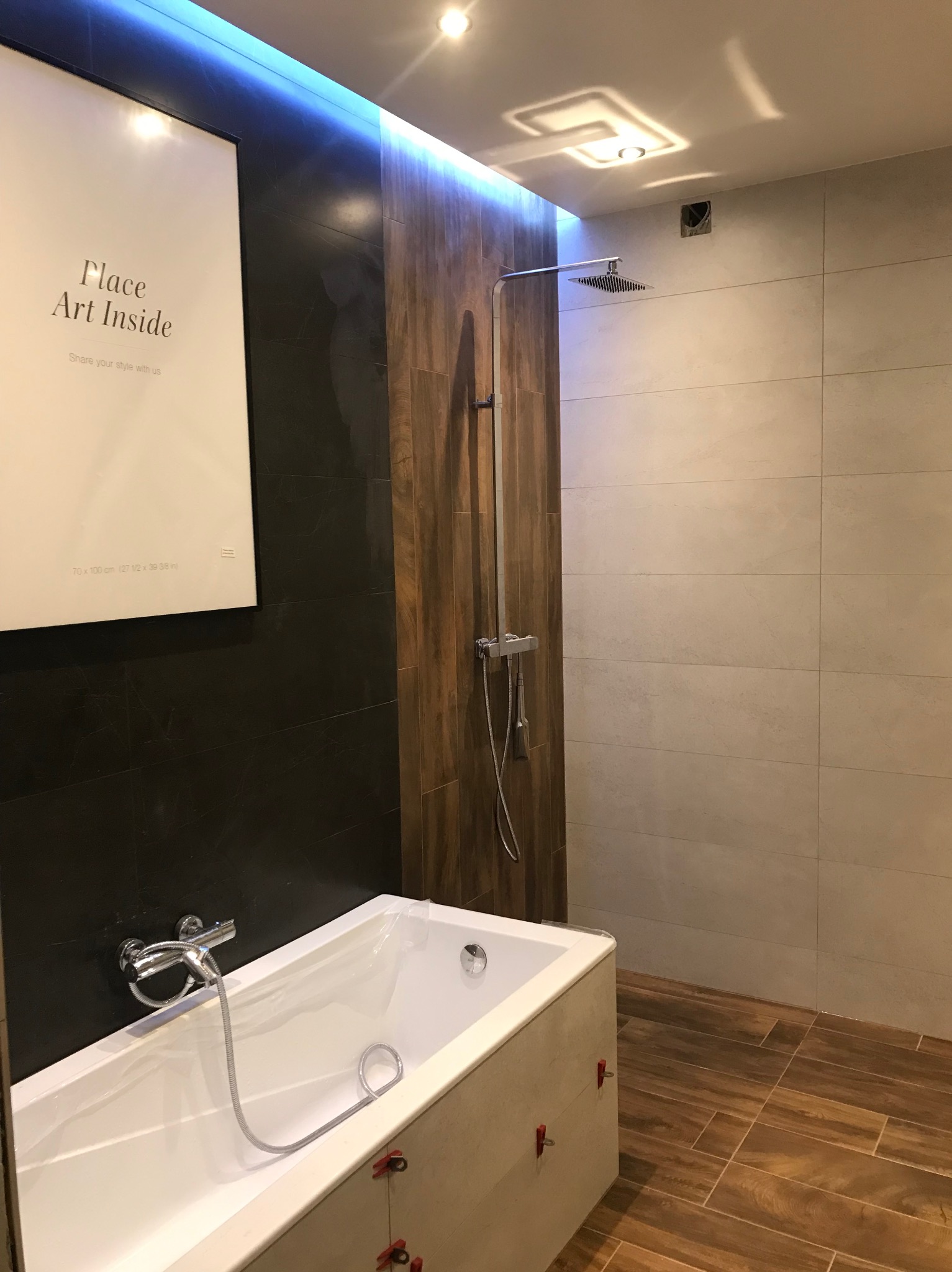 Nowoczesna łazienka z wanną obłożoną płytkami, panelami imitującymi drewno i deszczownicą; na ścianie plakat z napisem 'Place Art Inside'.
