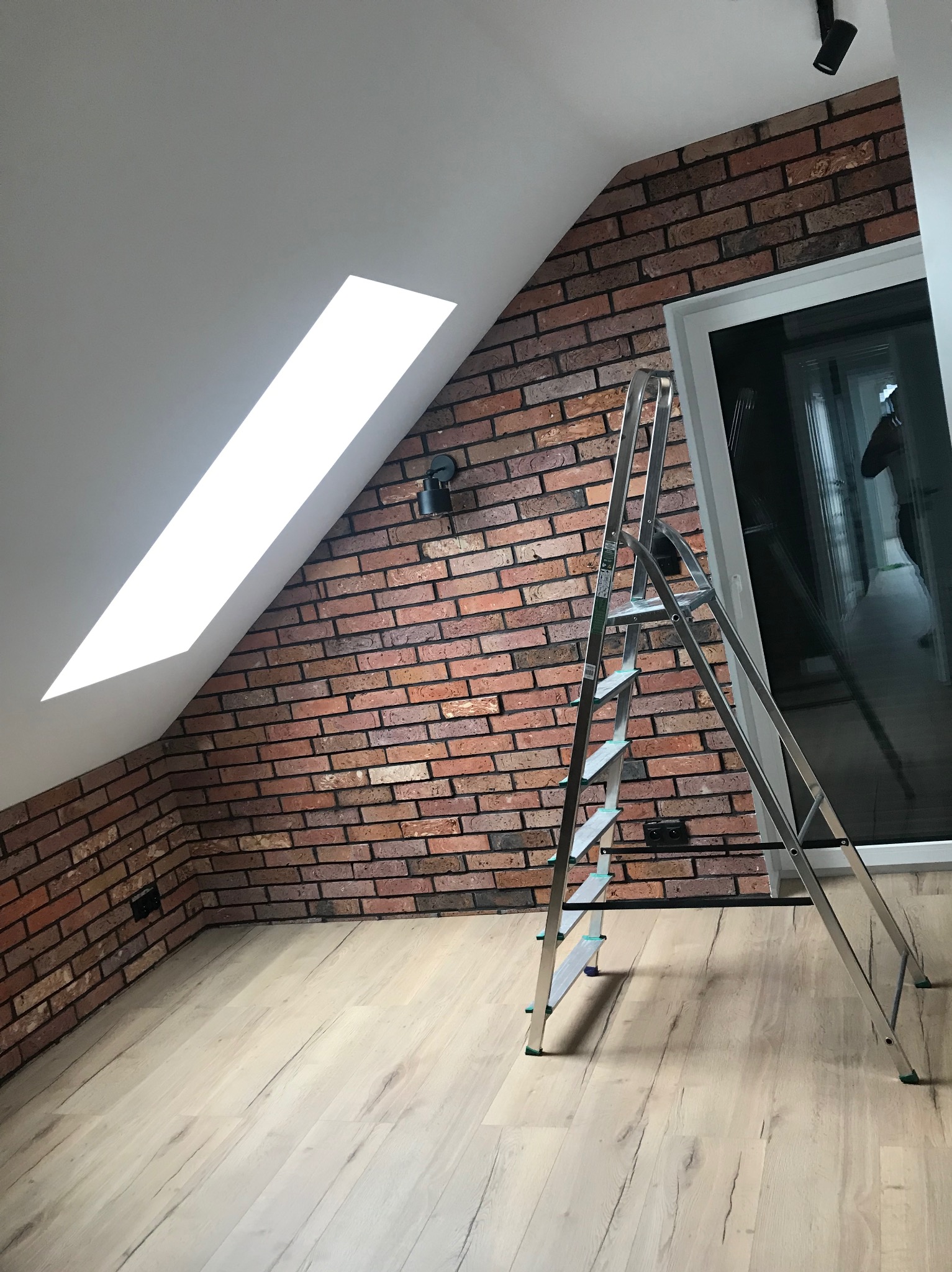 Poddasze po remoncie: ceglana ściana z oknem dachowym, aluminiowa drabina, jasna drewniana podłoga, czarne oświetlenie punktowe.