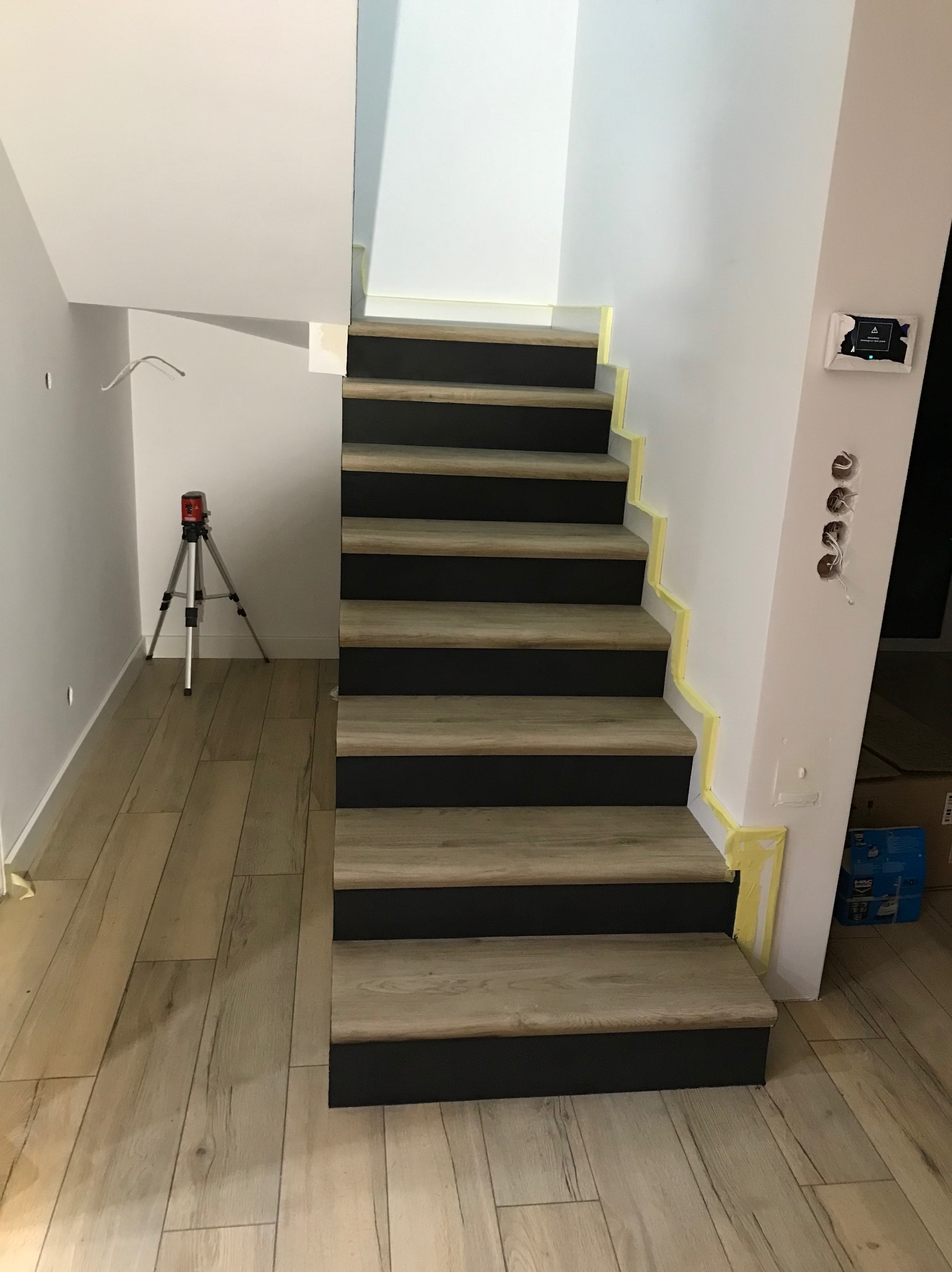 Schody z drewnopodobnymi stopniami i czarnymi podstopnicami, zabezpieczone taśmą malarską, z laserowym niwelatorem na trójnogu w tle oraz widocznymi otworami na gniazdka elektryczne.