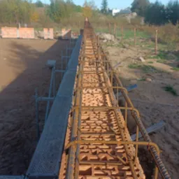 Wąska ściana z cegieł dziurawki zbrojona stalą, widok z góry wzdłuż ściany, rusztowanie po lewej stronie, w tle teren budowy i drzewa.