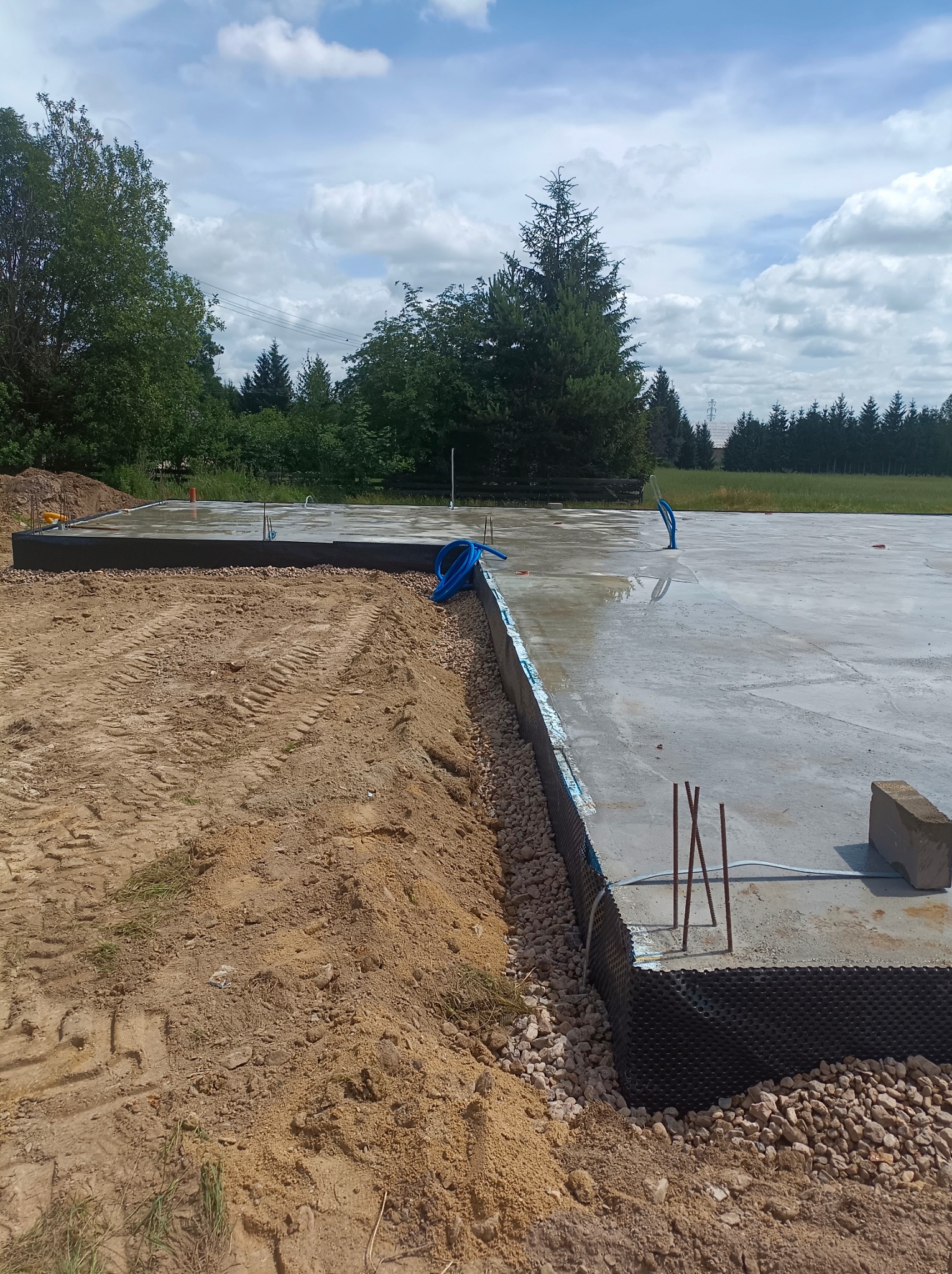 Wylany fundament z hydroizolacją i wystającymi prętami zbrojeniowymi, widoczne ślady opon na ziemi, błękitne rurki instalacyjne wystające z betonu.
