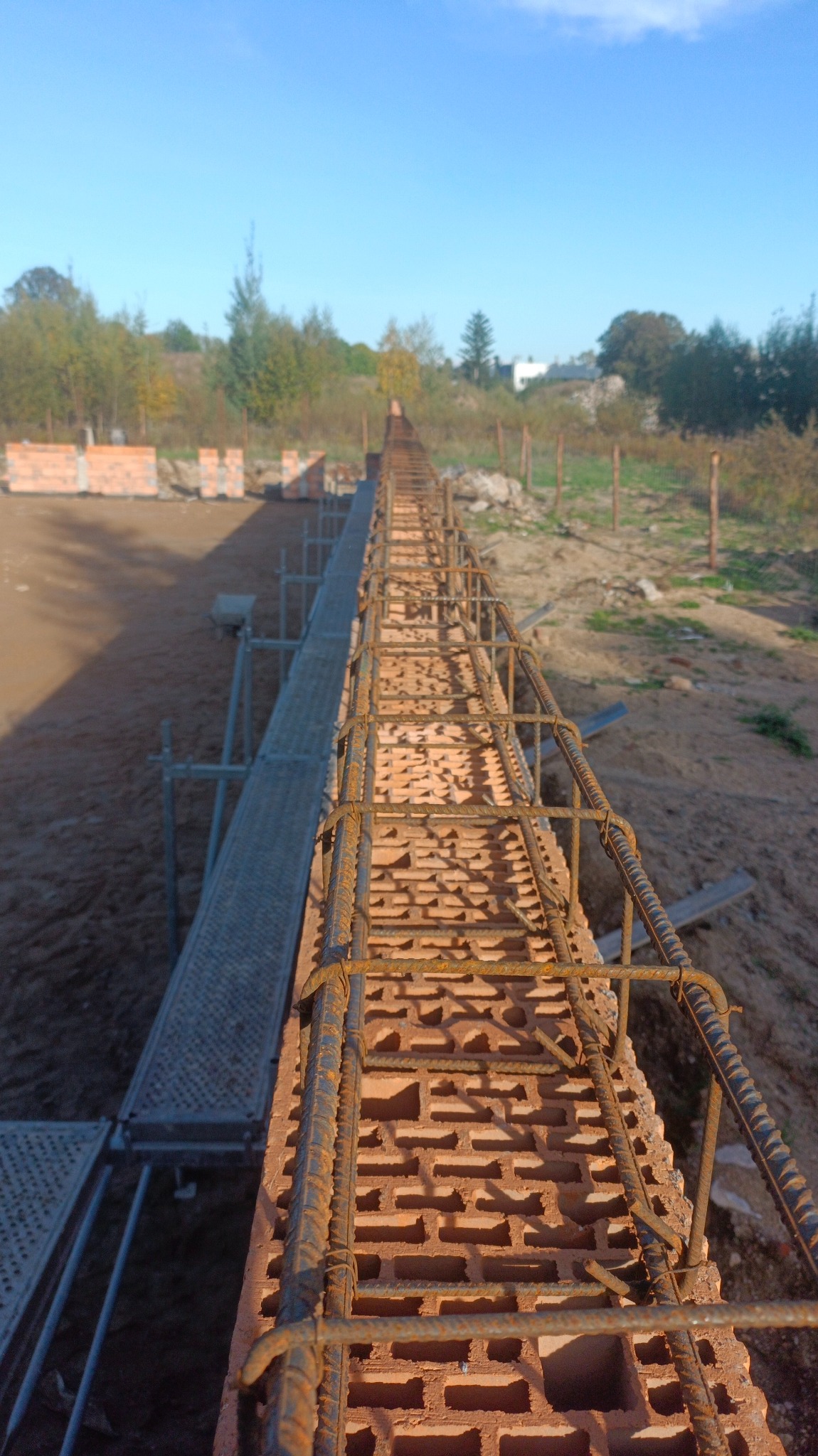 Wąska ściana z cegieł dziurawki zbrojona stalą, widok z góry wzdłuż ściany, rusztowanie po lewej stronie, w tle teren budowy i drzewa.