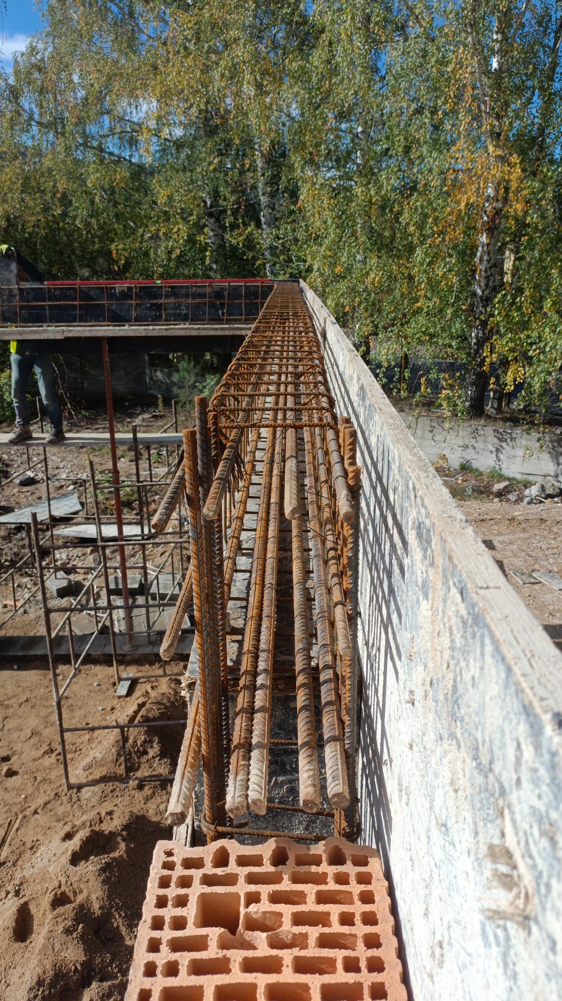 Zbrojenie w szalunku fundamentu z wystającymi prętami, widok z góry na pustak ceramiczny, w tle drzewa.