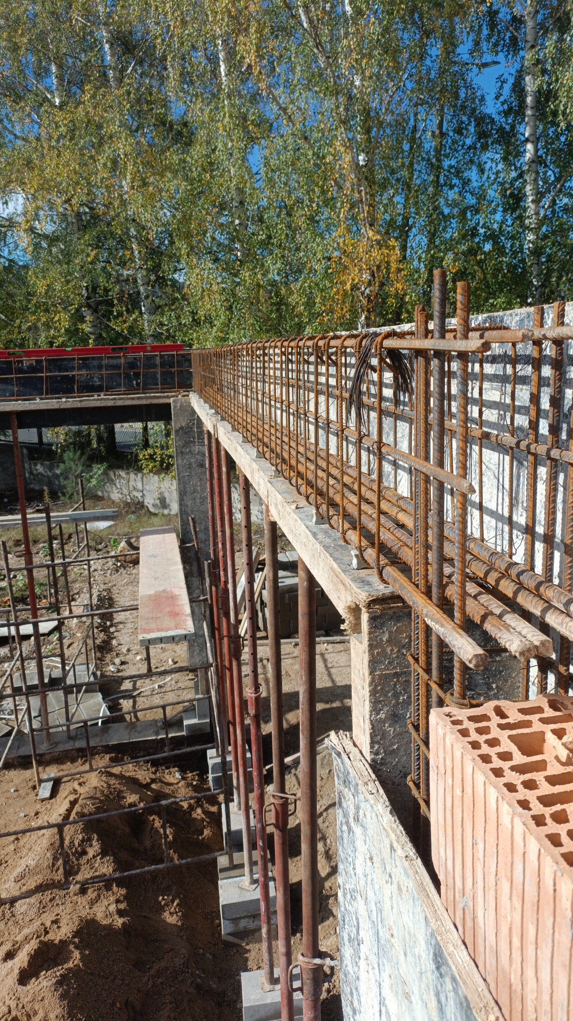 Wysoki kąt widzenia na szalunek zbrojeniowy pod przyszły strop, wsparty na metalowych stemplach, z widoczną cegłą dziurawką i drzewami w tle.